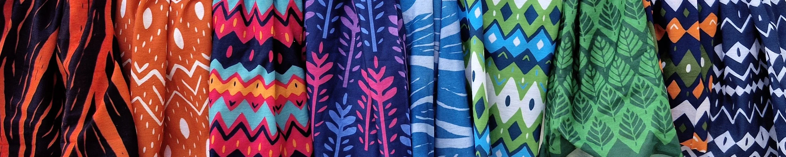 Various colorful fabrics hanging side by side, featuring different patterns including zigzags, stripes, abstract shapes, and leaf designs in a wide range of colors like orange, pink, blue, and green.