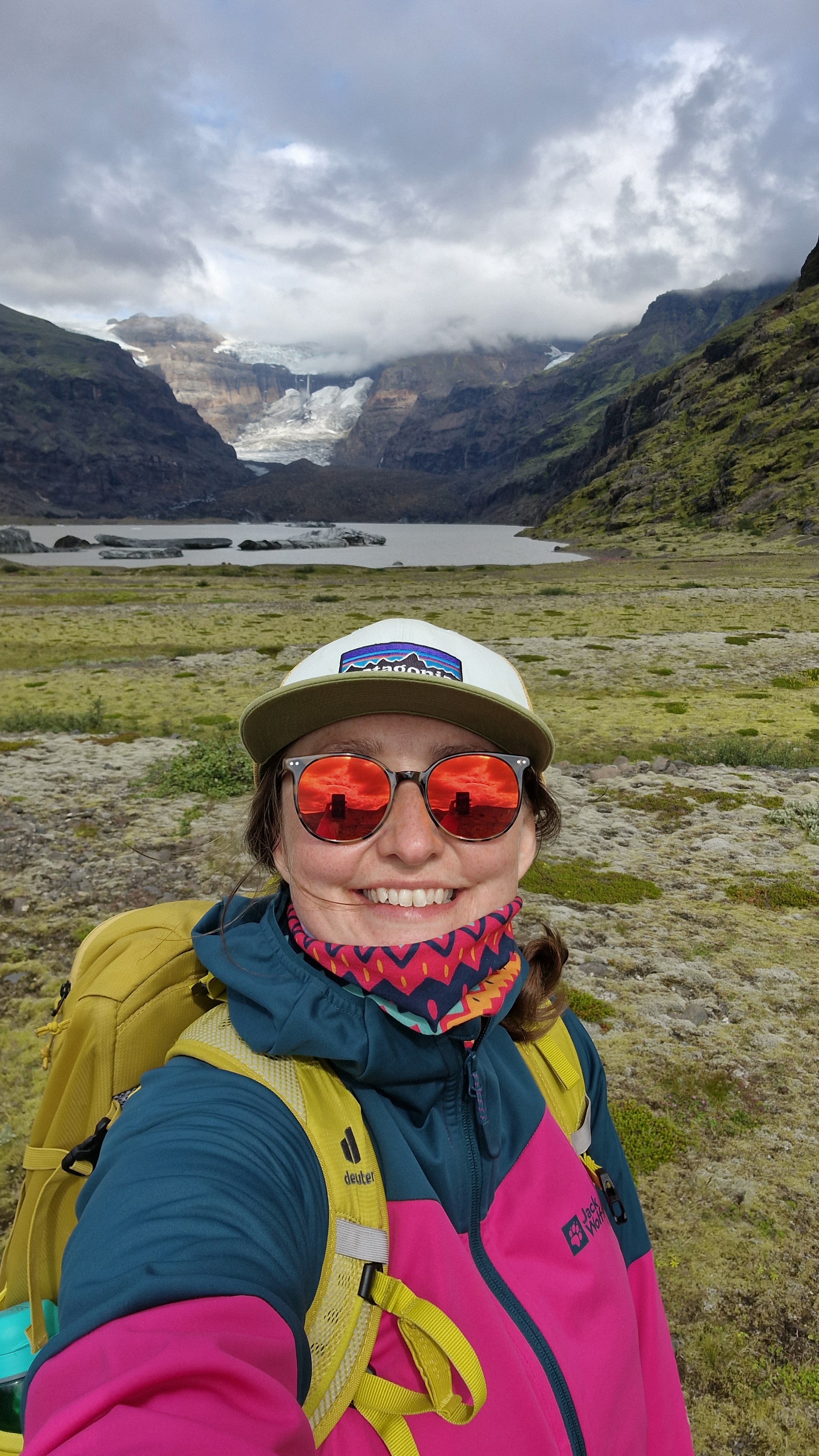 A smiling woman wearing sunglasses, a colorful neck warmer, a yellow backpack, and outdoor gear, standing in front of a scenic Icelandic landscape with mountains, a lake, and cloudy sky.