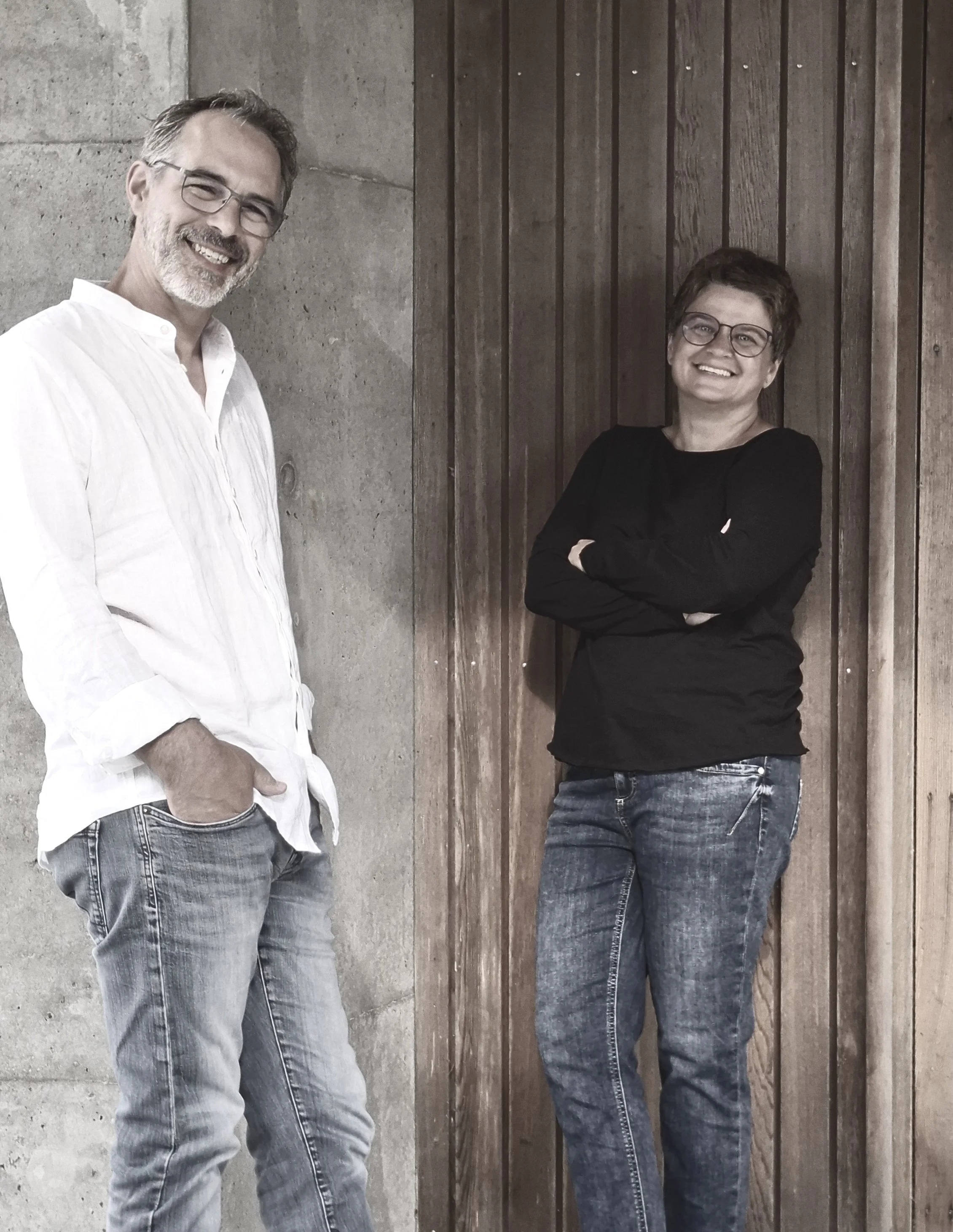 Ein Mann in weißem Hemd und eine Frau in schwarzem Pullover, stehen vor einer Wand aus Holz und Beton.