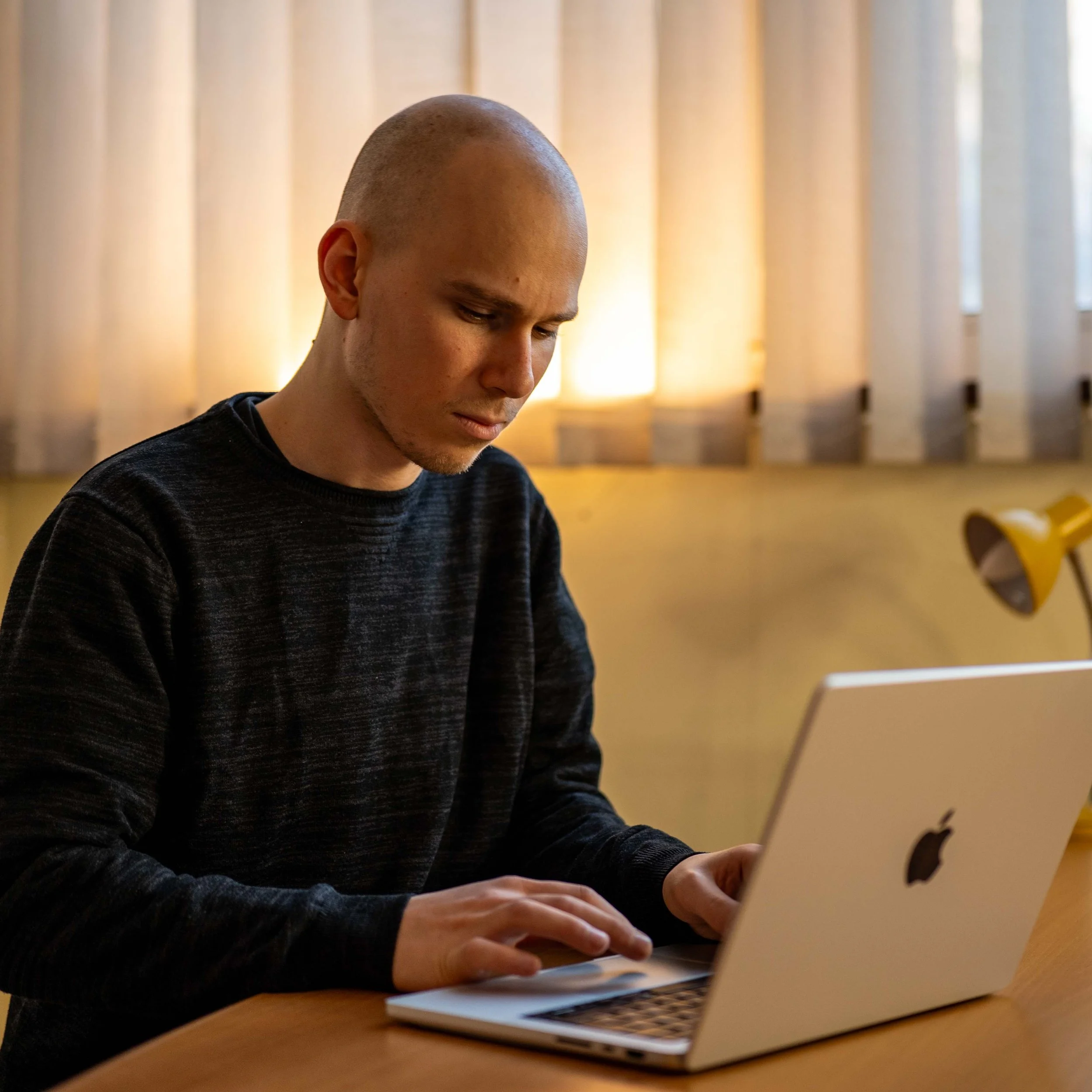 Person working on a laptop at a desk with a lamp nearby and curtains in the background.