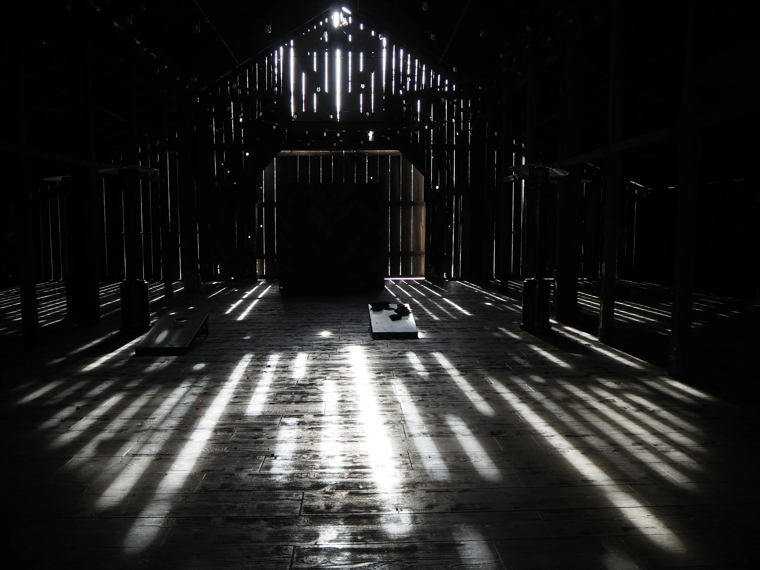 Interior of a dark, empty barn with sunlight streaming through gaps in the wooden walls, casting shadows on the floor.