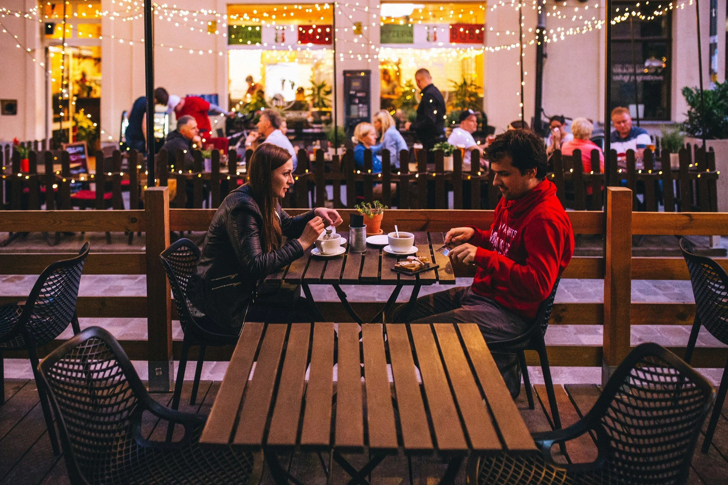 Outdoor restaurant seating with two people dining, tables, chairs, and string lights overhead.