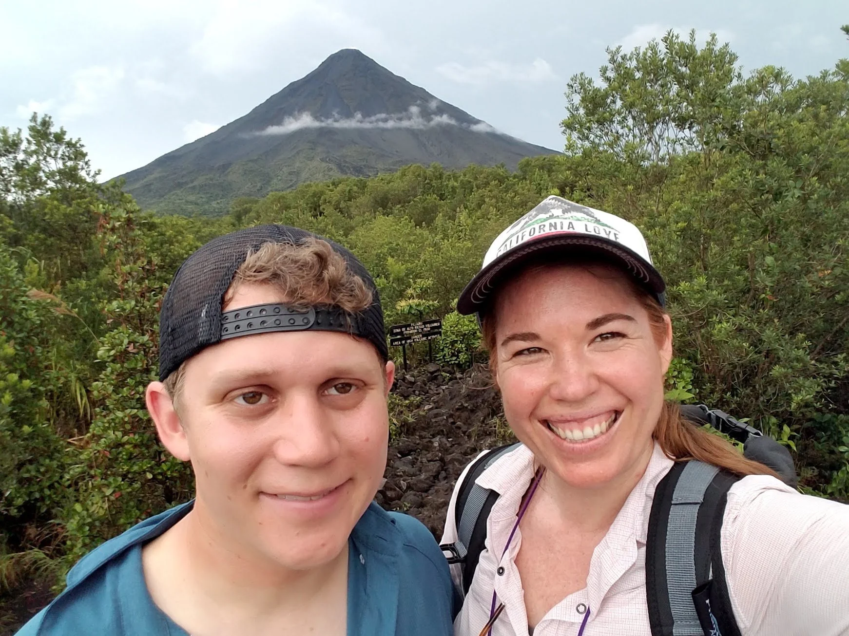 Couple with backpacks smiling in front of Arenal Volcano in Costa Rica during a hiking adventure.