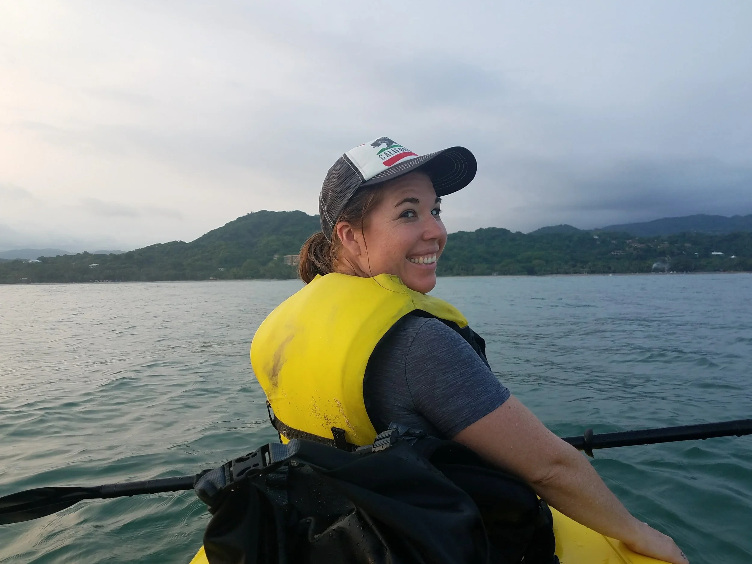Travel advisor kayaking in Costa Rica at dusk, wearing a life jacket and smiling back at the camera, with lush green hills along the water.