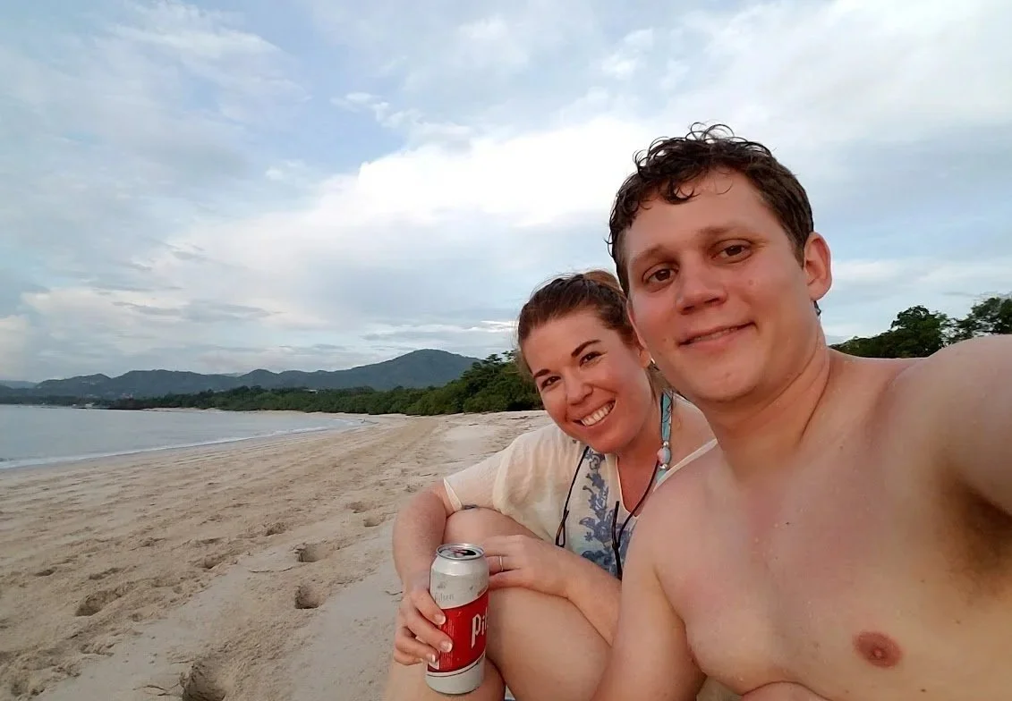 Couple smiling in a selfie on the sand at Playa Minas near Tamarindo, with calm water and green hills behind them.