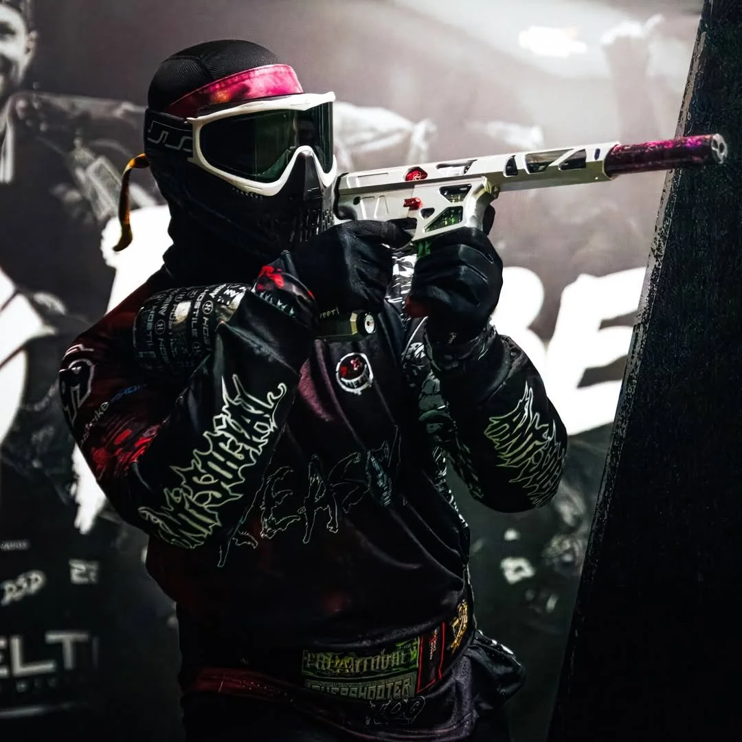 A person wearing a black helmet with pink and white accents, goggles, all black gloves, and a black jacket with patches, holding a pink and white paintball gun.