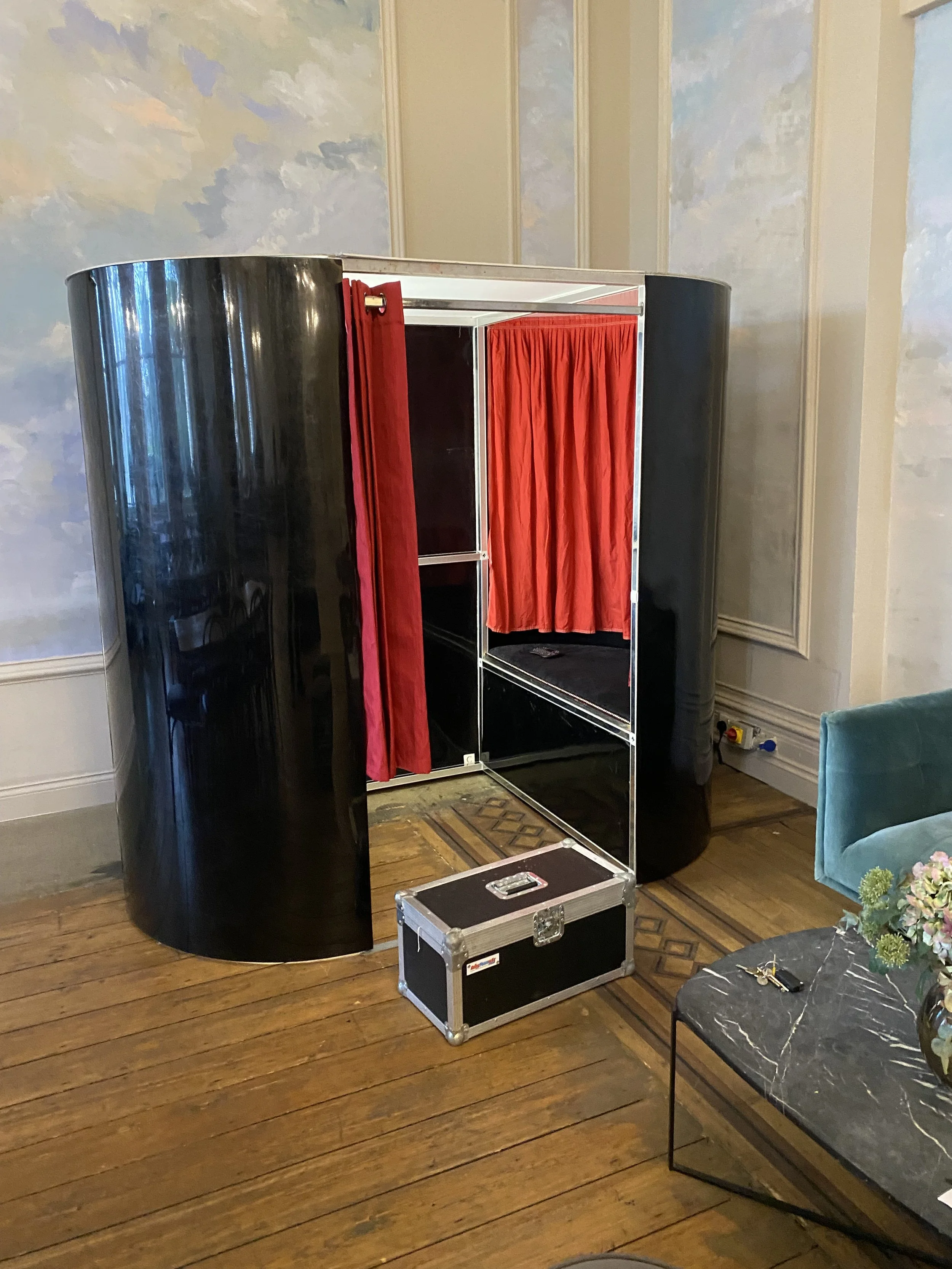 A black and silver photo booth with red curtains inside, placed on a wooden floor in a room with ornate wall paneling and a painted sky mural.