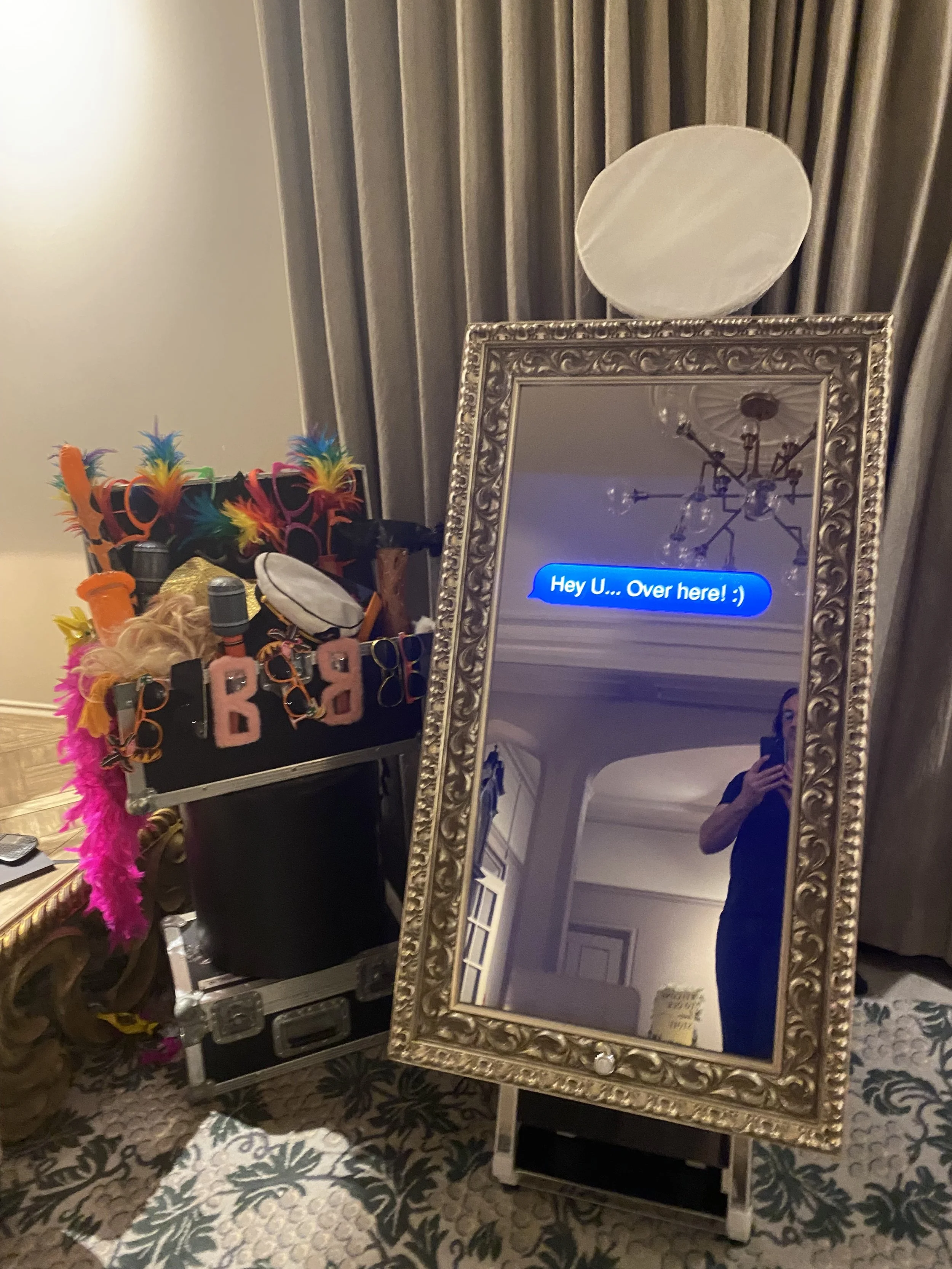 A decorative mirror with an ornate gold frame on a stand, reflecting an interior room and a person taking a photo. The mirror has a blue text message overlay that reads 'Hey U... Over here! :)'. Next to the mirror is a Black storage case filled with colorful feather boas, costume jewelry, sunglasses, and accessories.
