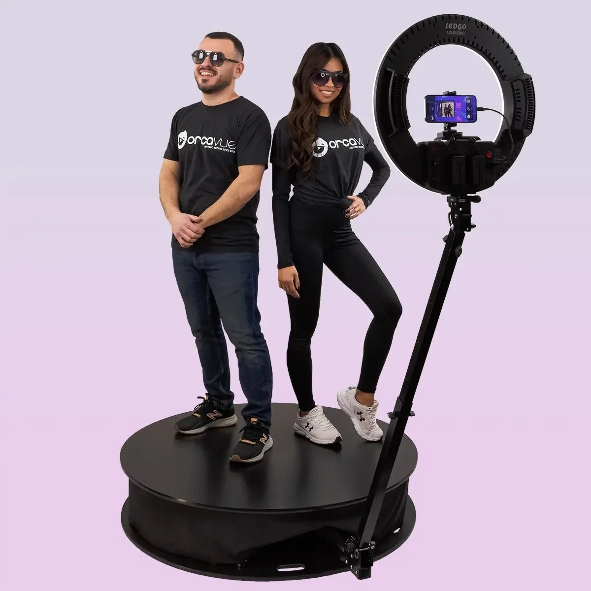 Two people, a man and a woman, standing on a circular platform in front of a ring light and a smartphone mounted for recording, both wearing black shirts with white logos and sunglasses.
