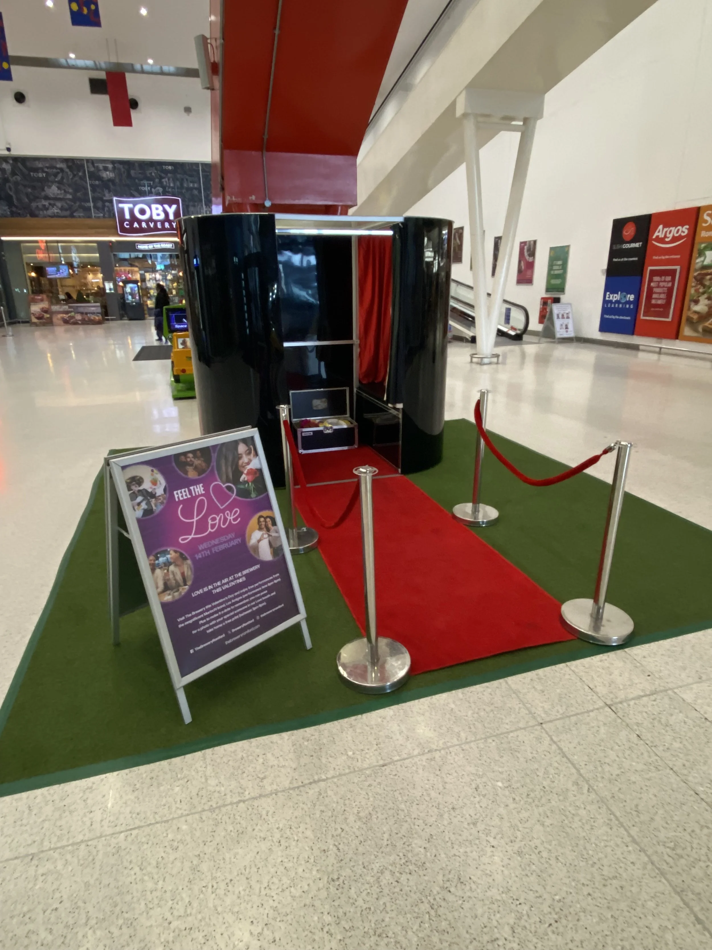A photo of a small black photo booth with a red carpet and velvet curtain inside, set up on green artificial turf in a shopping mall. A signboard nearby promotes a Valentine's Day event.