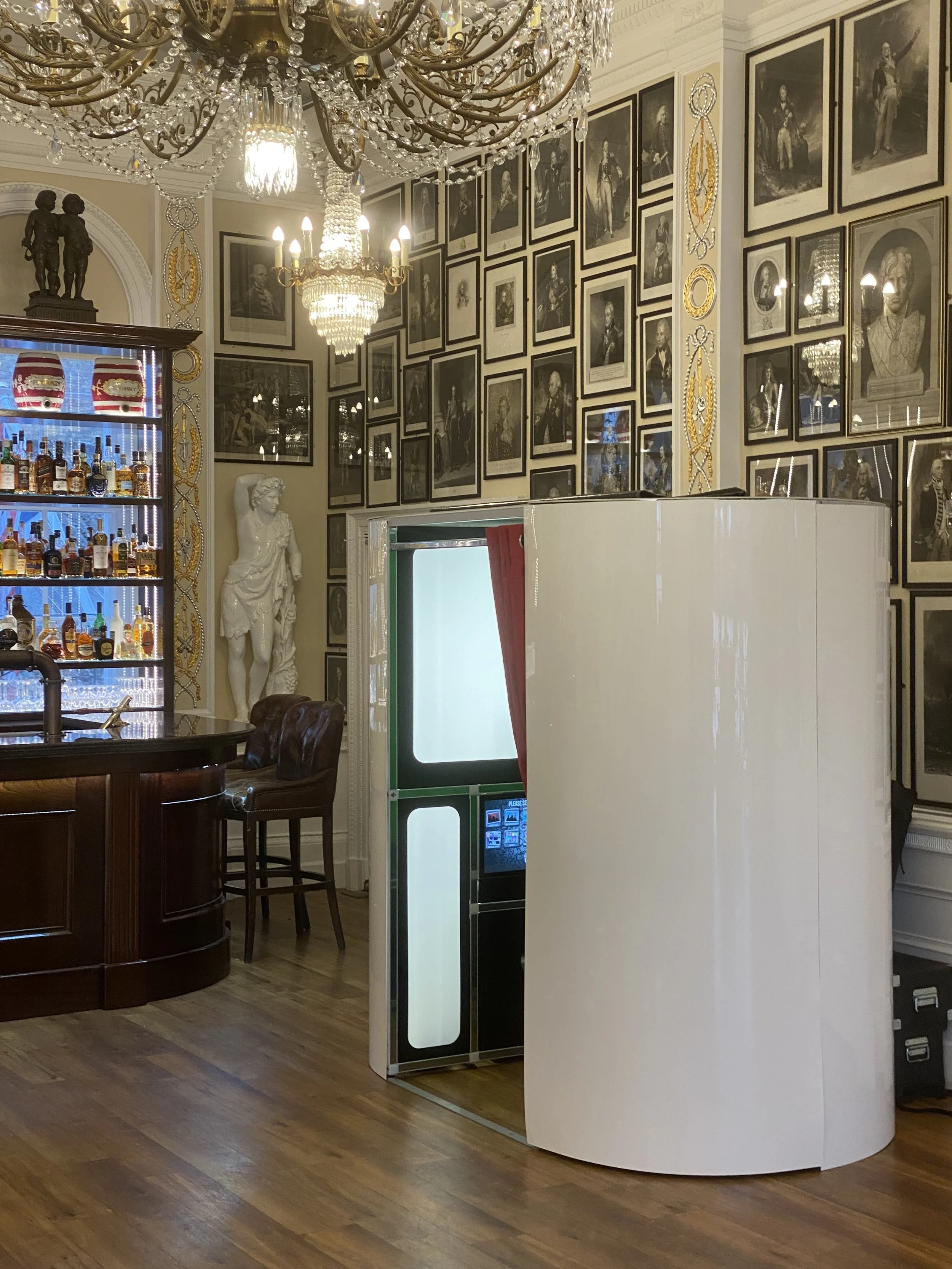 Elegant room with chandeliers, framed black-and-white portraits on the walls, a bar area with bottles, a statue, and a photo booth.