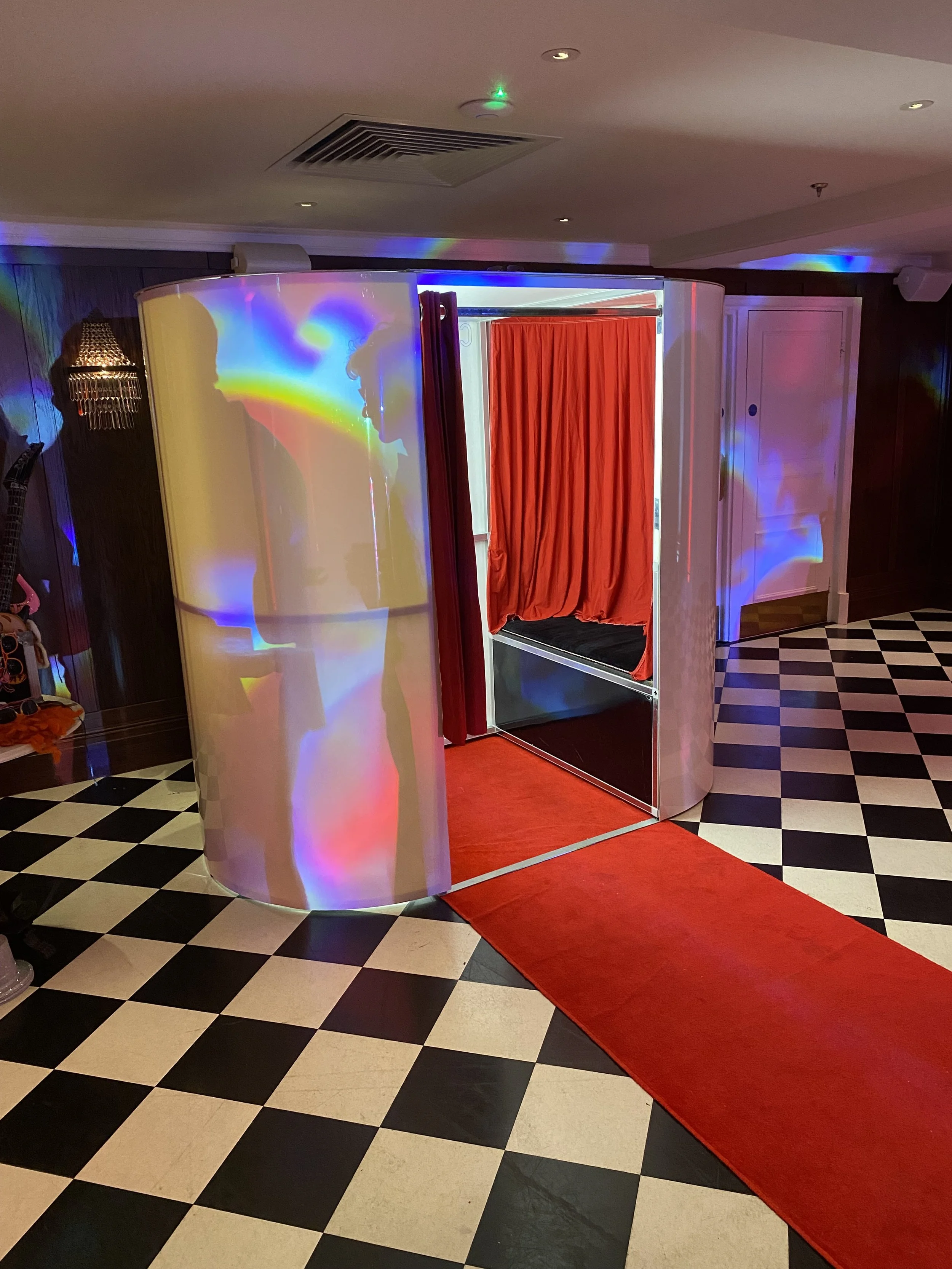 Photo of a photo booth with red curtain, illuminated by colorful lights, and a checkered black and white floor.
