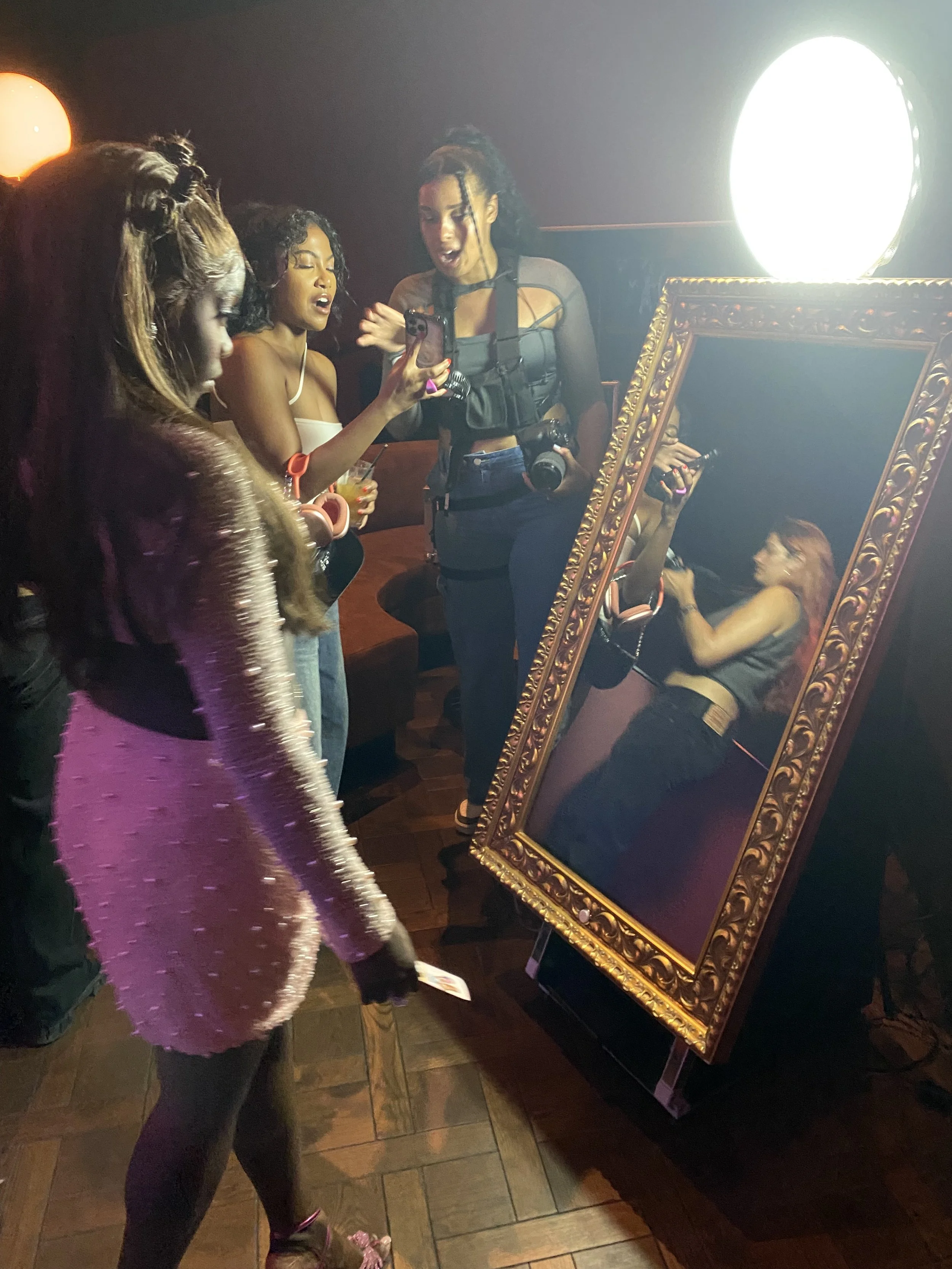 Group of women taking a selfie in front of a large, ornate mirror at a dimly lit venue with a bright light in the background.