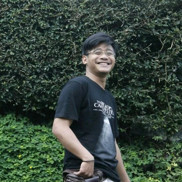 Young man smiling outdoors with a lush green hedge in the background, wearing glasses and a black T-shirt.