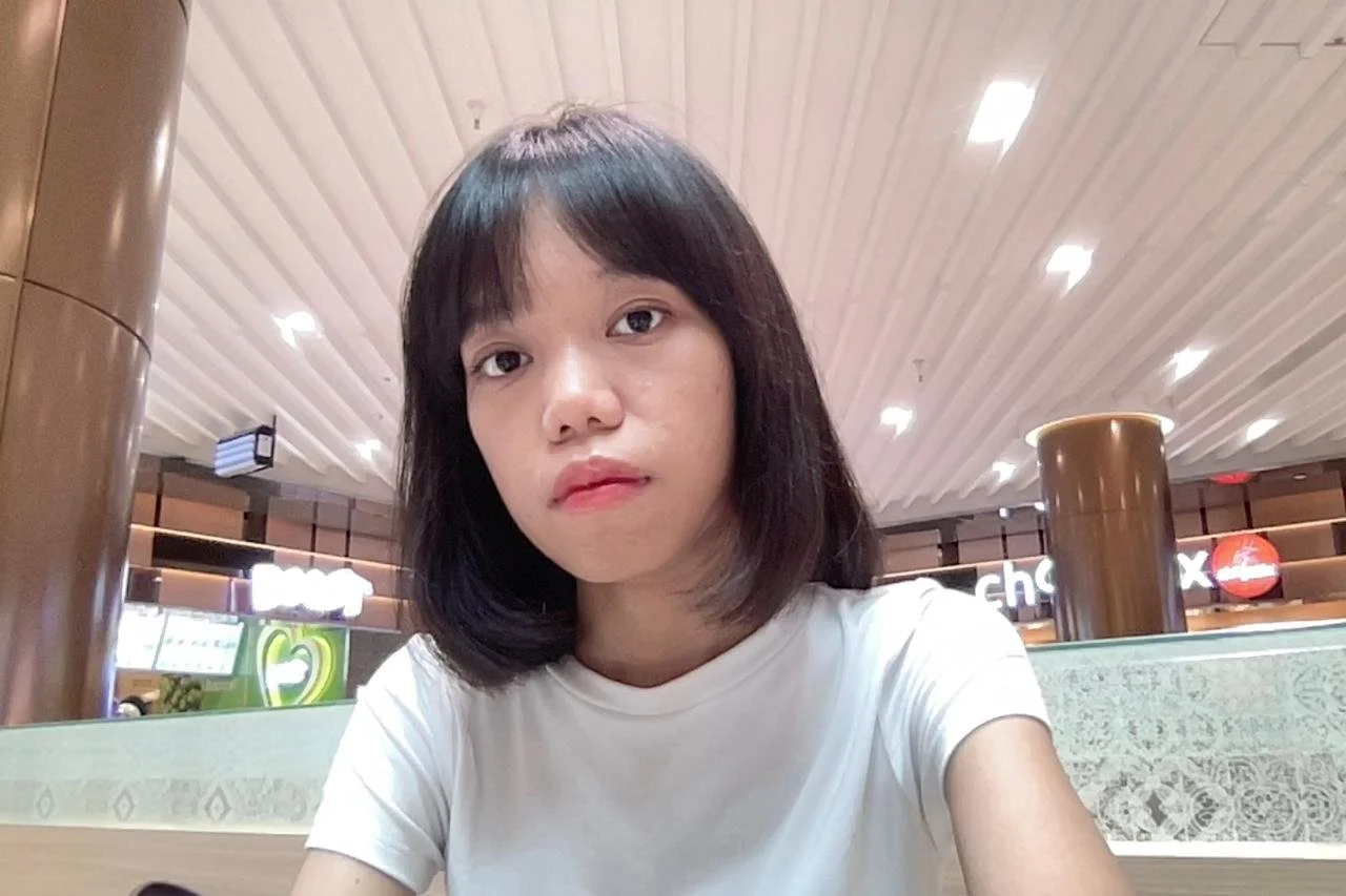 A young woman with shoulder-length black hair and a white T-shirt sitting inside a shopping mall or food court.