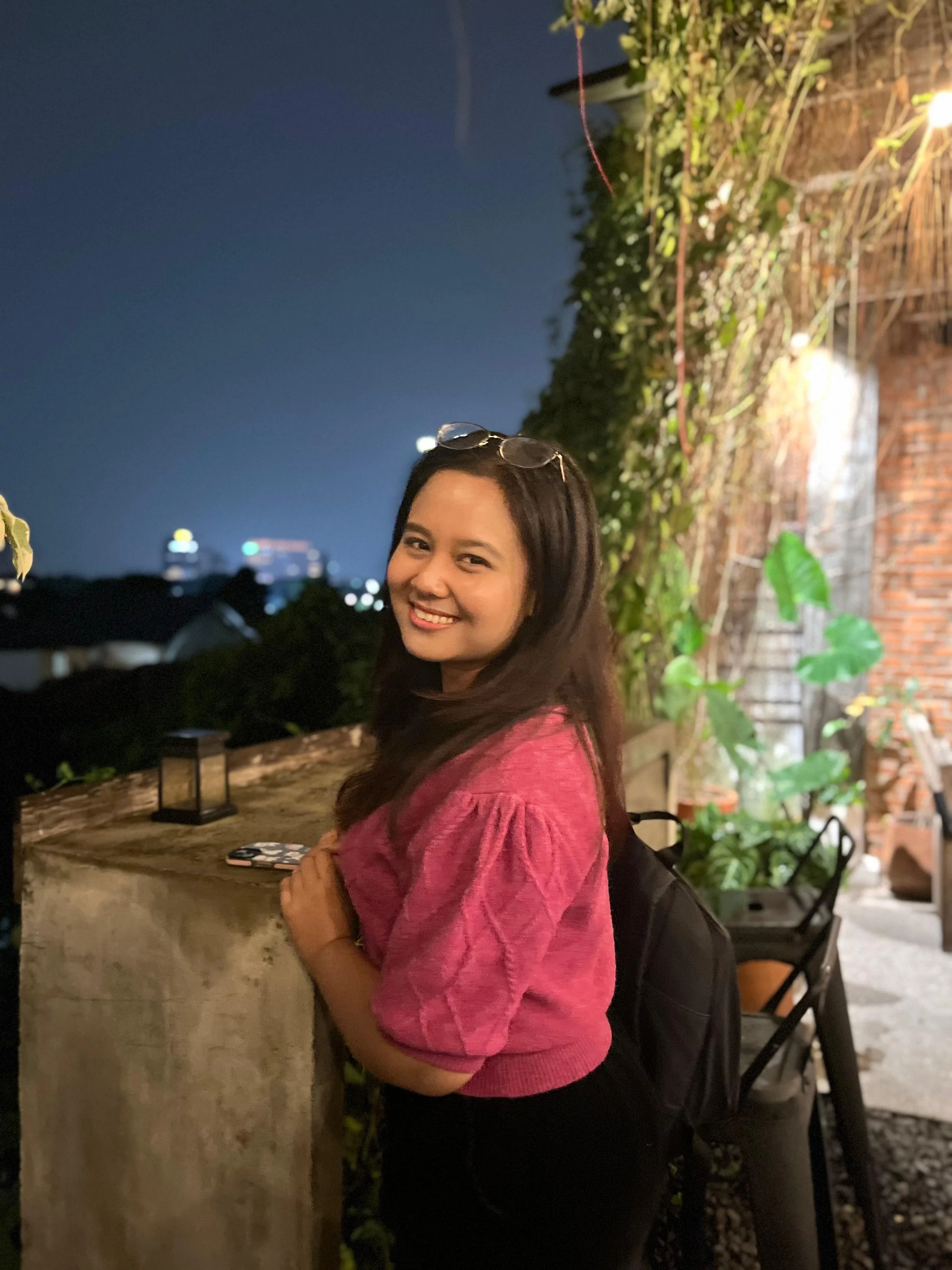 A smiling woman with glasses on her head wearing a pink sweater and black pants, standing outdoors at night with city lights in the background, leaning on a concrete ledge with a lantern and a phone on top, surrounded by greenery and brick wall.