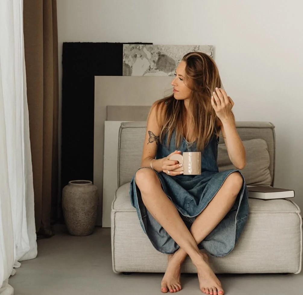 A woman with brown hair and a tattoo on her left arm sitting on a light-colored sofa, holding a beige mug, looking towards her left. She is wearing a sleeveless denim dress and is barefoot. There are framed canvases and a large vase in the background, and a book along with a closed notebook are placed beside her on the sofa.