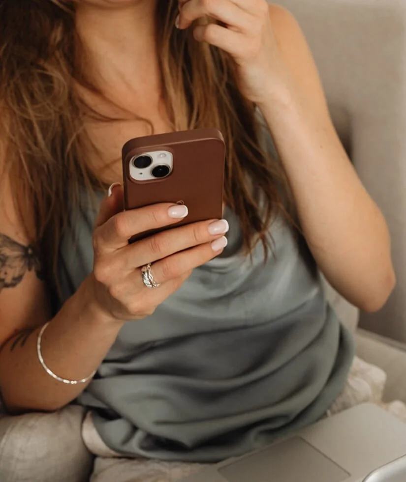 A woman with long, wavy red hair is sitting on a couch, holding a smartphone in her right hand, taking a selfie. She is wearing a gray sleeveless top, a ring on her finger, a bracelet, and has a tattoo on her upper left arm. The background shows part of the couch and a neutral-colored wall.