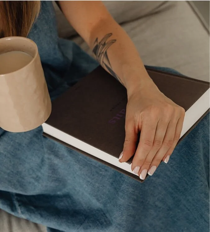 Close-up of a person's hand resting on an open notebook, with a tattoo of abstract lines visible on their forearm, while holding a mug.
