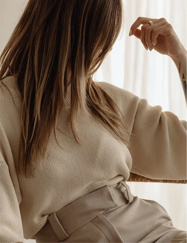 A woman with long, brown hair wearing a beige sweater and matching high-waisted pants, sitting near a window with beige curtains.