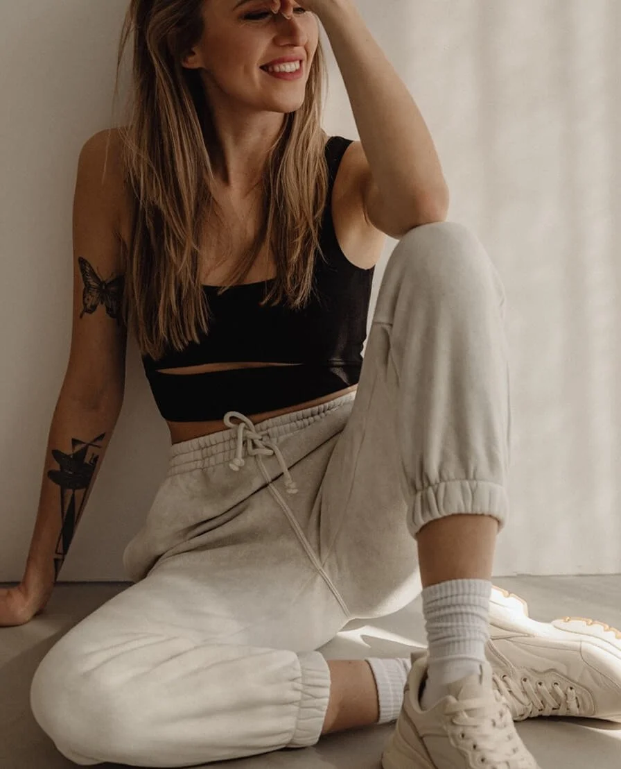 A young woman sitting on the floor, smiling, wearing a black crop top, beige sweatpants, white socks, and sneakers, with tattoos on her arms and long brown hair.