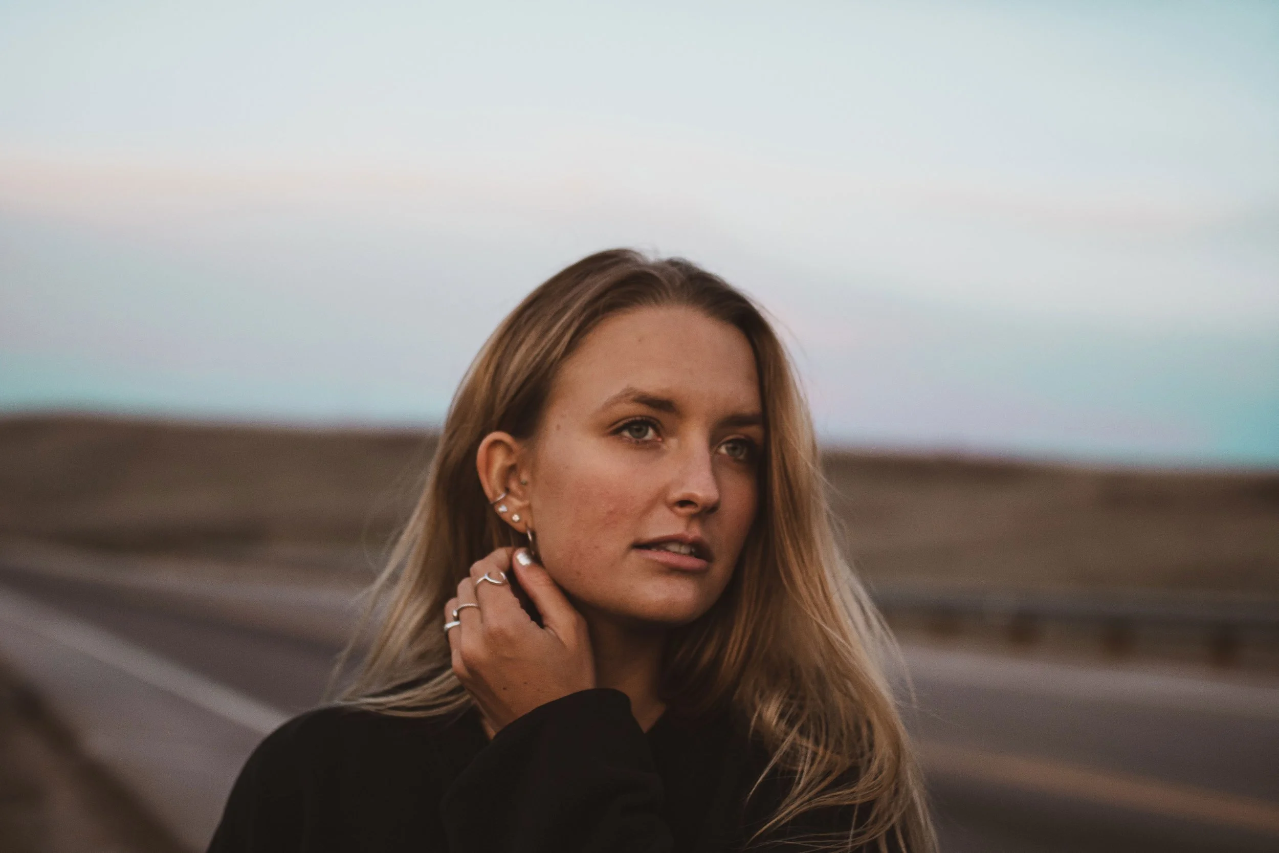 A young woman with long blonde hair, multiple earrings, and rings on her fingers, stands outdoors in a rural area with a road and hills in the background during the daytime, with a neutral expression.