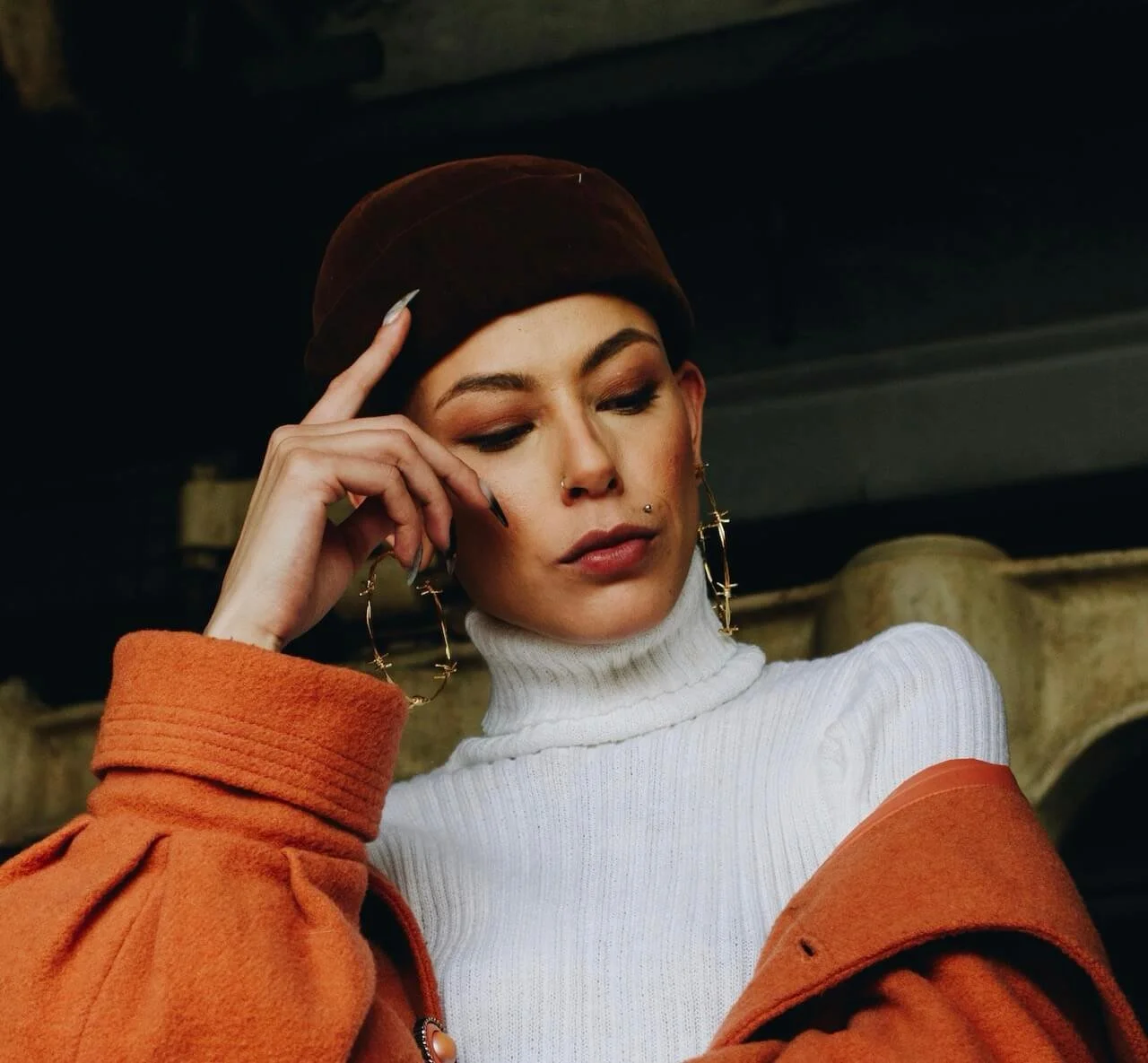 A young woman with multiple facial piercings and large hoop earrings, wearing a brown beanie, white turtleneck, and orange coat, looking down thoughtfully with her hand near her temple.