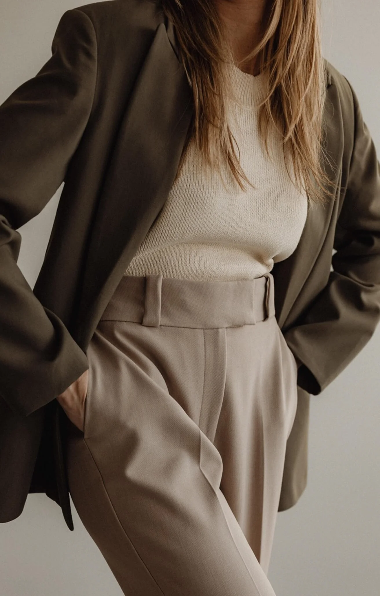 A person wearing beige high-waisted trousers, a cream sweater, and an olive-green blazer.