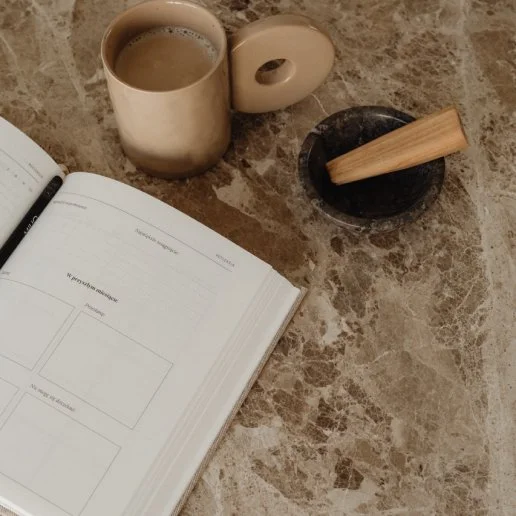A coffee mug filled with coffee, a black mortar and pestle with a wooden pestle, an open notebook with a pen, all on a marble tabletop.