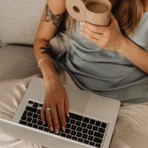 A woman sitting on a couch with a laptop on her lap, holding a coffee mug near her face, showing tattoos on her arm, a ring, and wearing a silvery top.