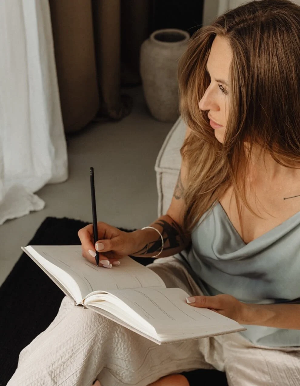 A woman with long brown hair and tattoos on her arm is writing in a notebook with a black pen, sitting on a bed or sofa in a room with beige and neutral decor.