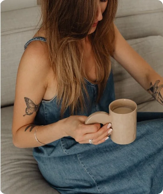 A woman with long hair and tattoos on her arms, wearing a denim dress, is sitting on a sofa holding a beige mug filled with a beverage.
