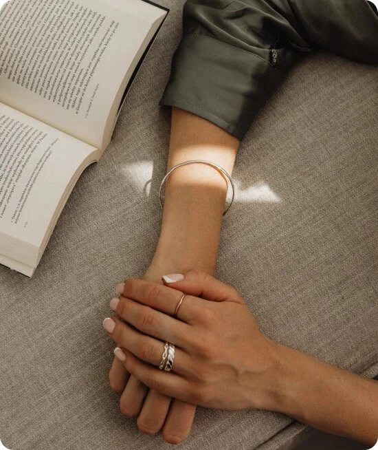 Two hands holding each other, one with rings and a bracelet, resting next to an open book on a sofa or cushion.