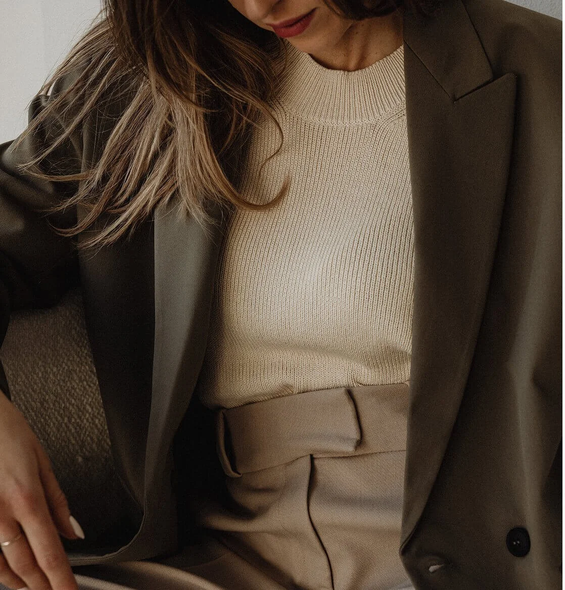 A woman wearing a beige knitted sweater, tan trousers, and a dark blazer with her shoulder-length hair partially over her face, sitting on a beige carpeted floor.