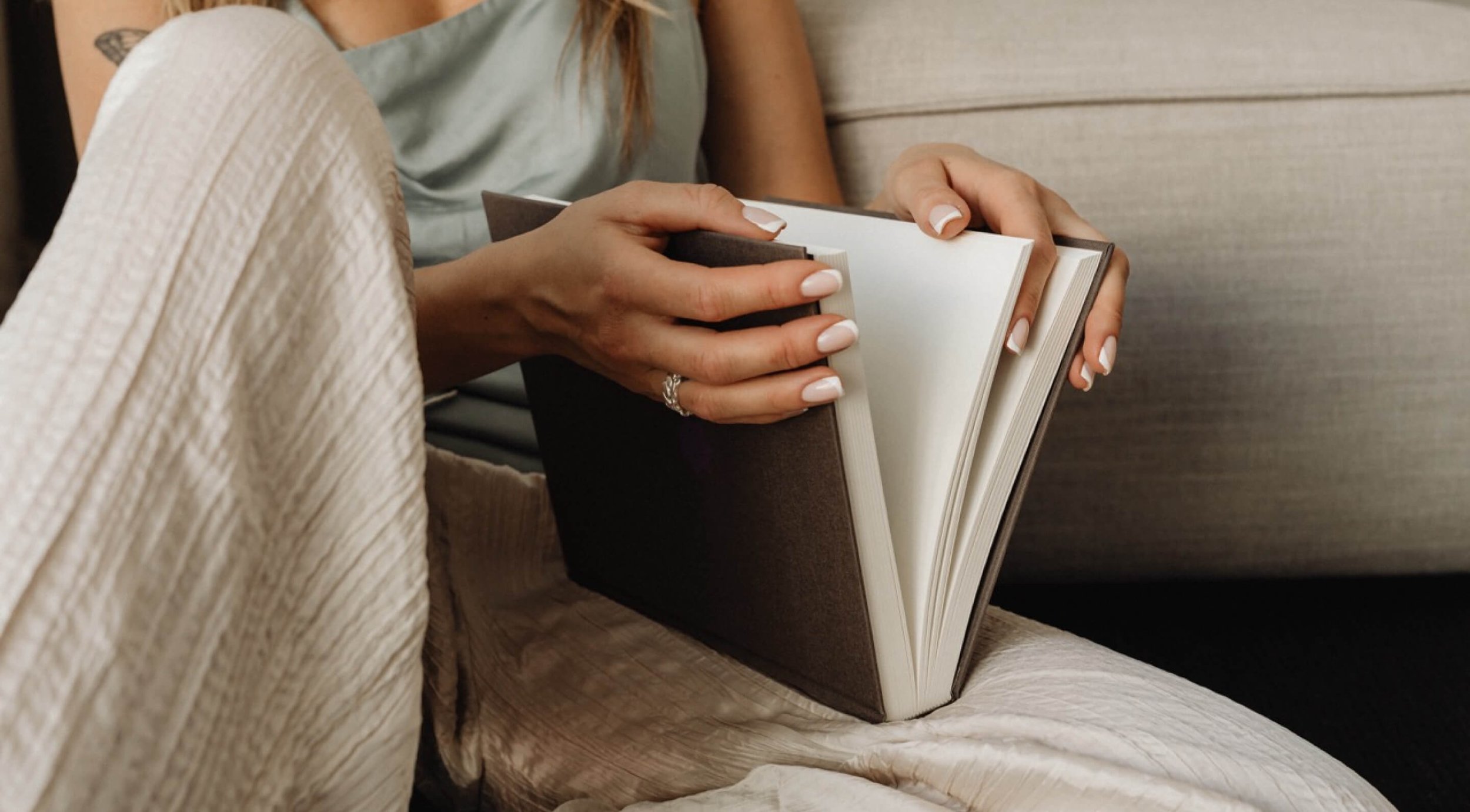 Person holding an open hardcover book with blank pages, sitting on a couch with legs crossed.