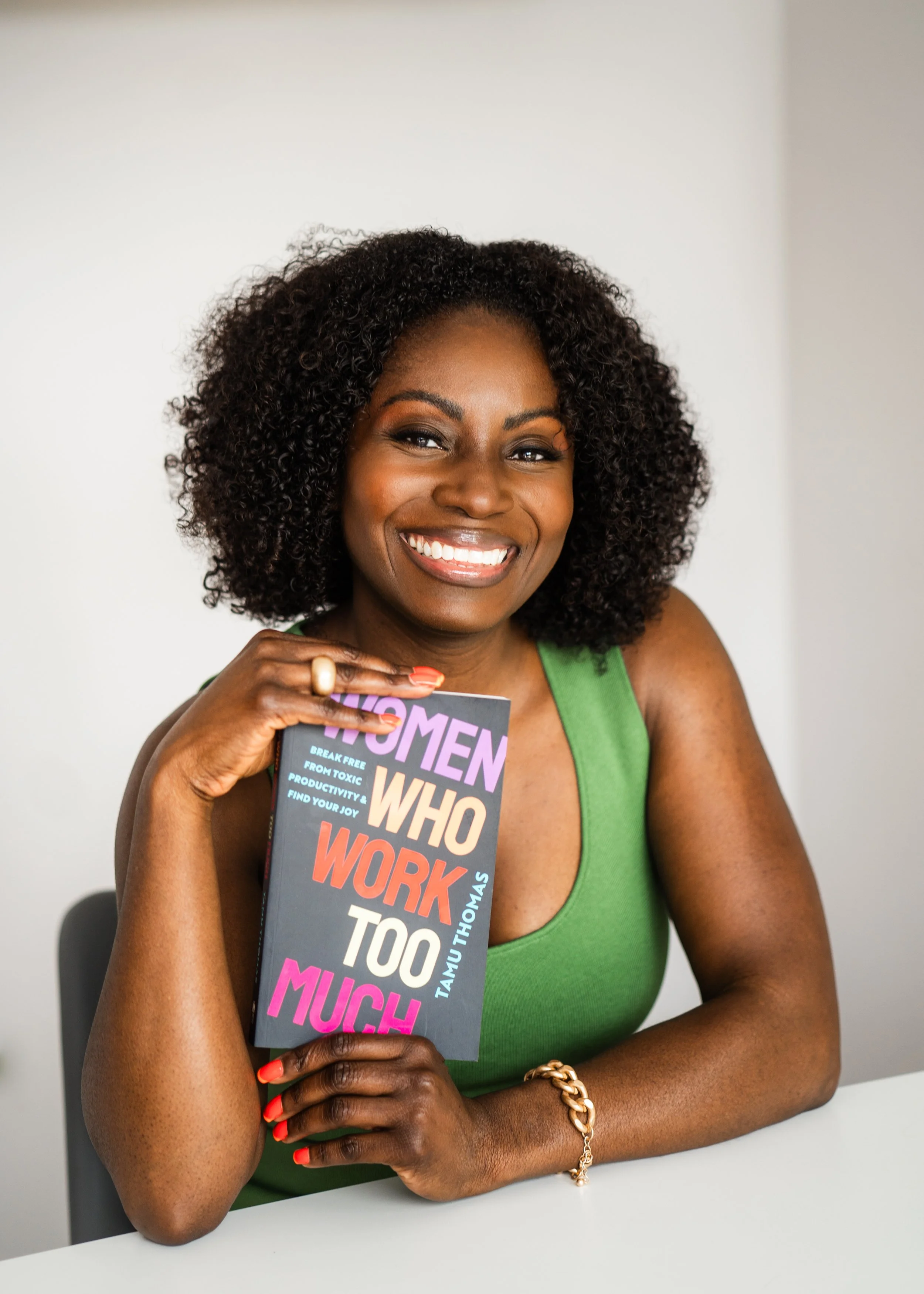 A smiling Black woman with curly hair, wearing a green sleeveless top and gold jewelry, holding a book titled 'Women Who Work Too Much' by TamU Thomas, sitting at a white table in a neutral room.