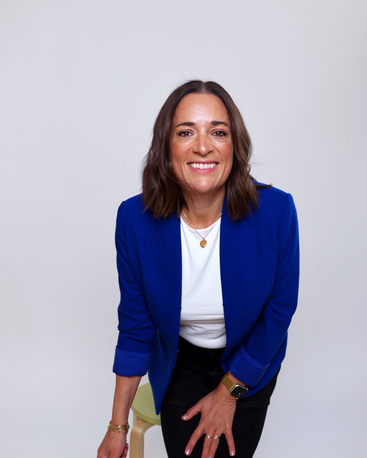 Woman with shoulder-length brown hair, smiling, wearing a blue blazer over a white top, black pants, a gold wristwatch, and a gold necklace with a heart pendant, sitting on a light-colored stool against a plain light gray background.