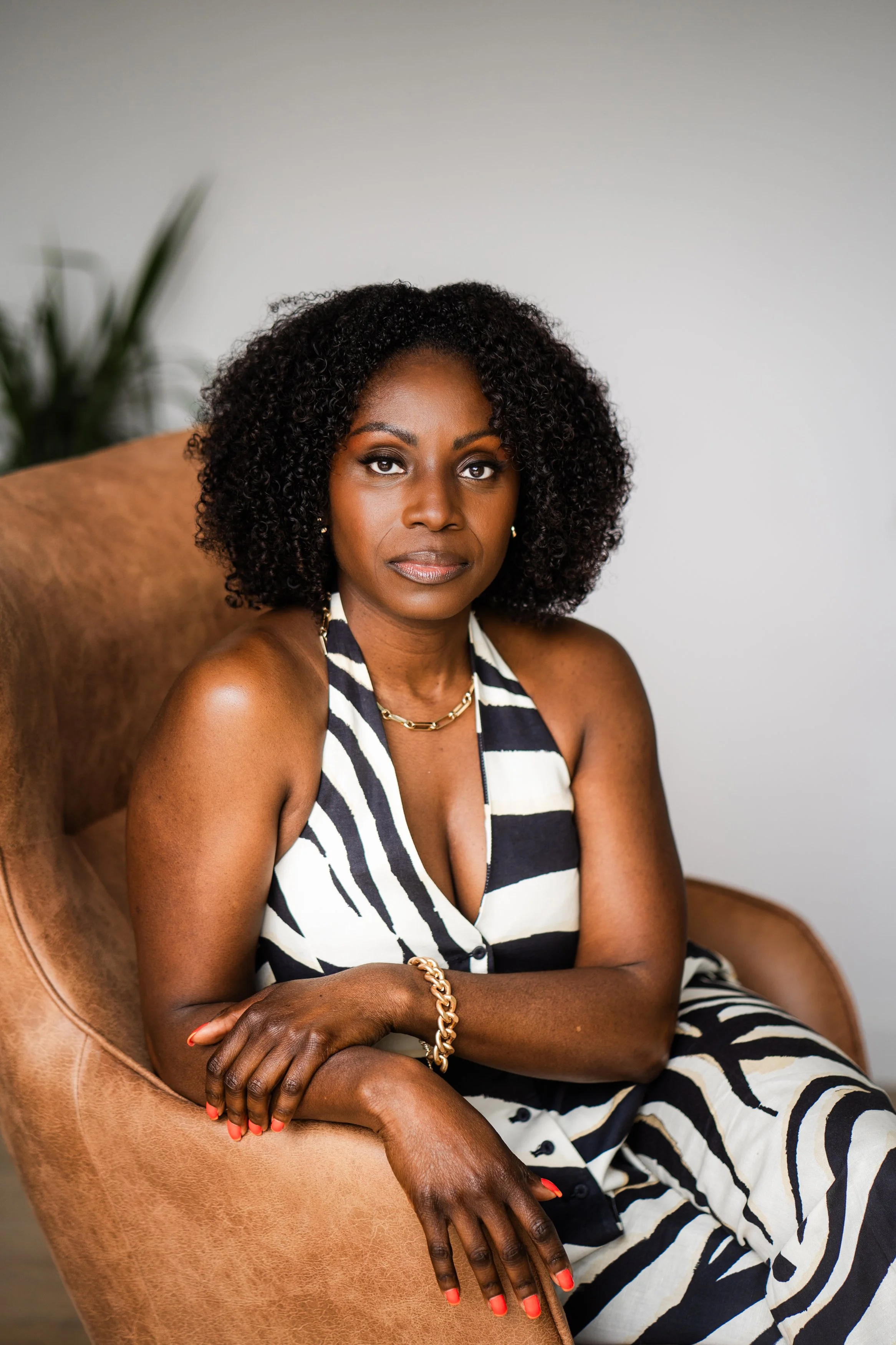 A woman with natural curly black hair sitting on a tan leather armchair, wearing a black and white zebra print sleeveless dress and gold jewelry, looking at the camera with a neutral expression.