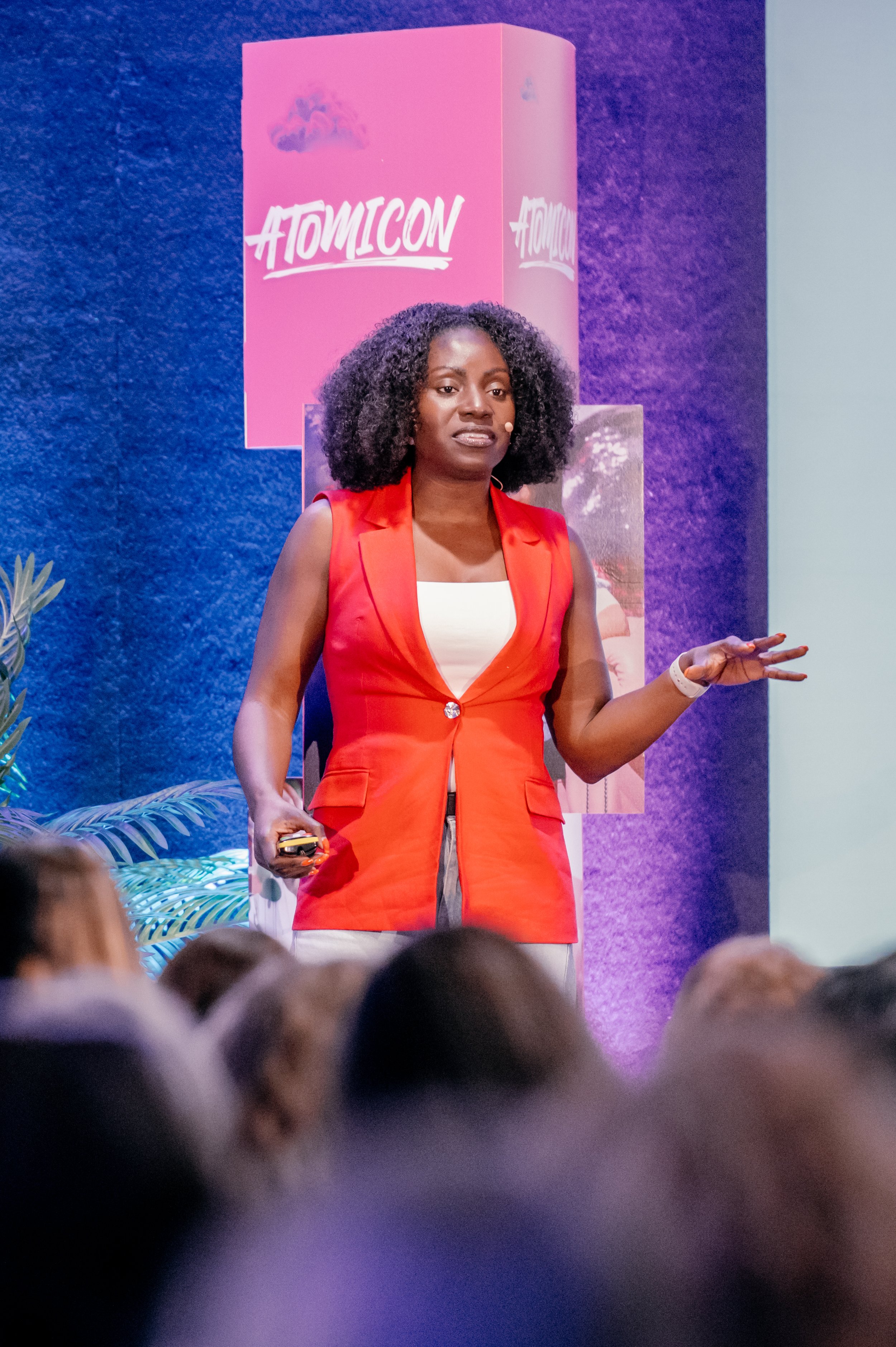 A woman giving a presentation at Atomicon conference, standing in front of a purple backdrop with Atomicon logo, dressed in a red sleeveless blazer over white top, with an audience in foreground.