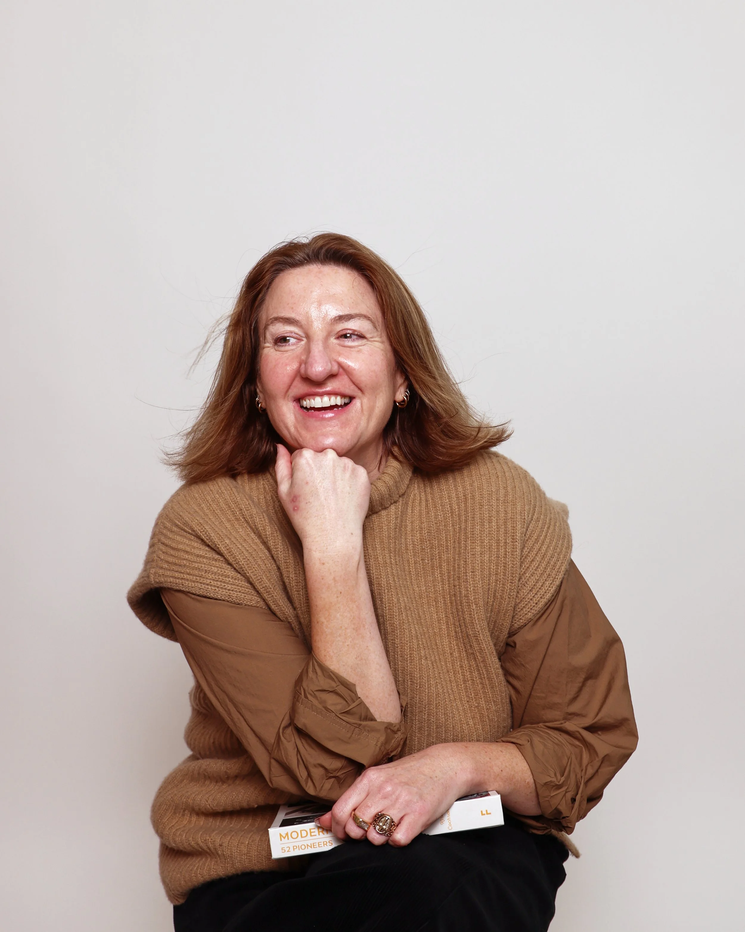 A woman with curly red hair, wearing a brown sweater, sitting against a plain white background, smiling and holding a brochure.