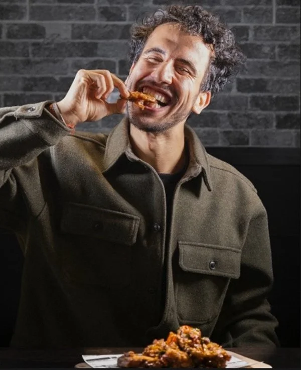 Ein lachender Mann mit dunklem Haar und Bart isst ein Stück Grillhähnchen in einer dunklen Umgebung mit Ziegelwand @ Wismar marketing