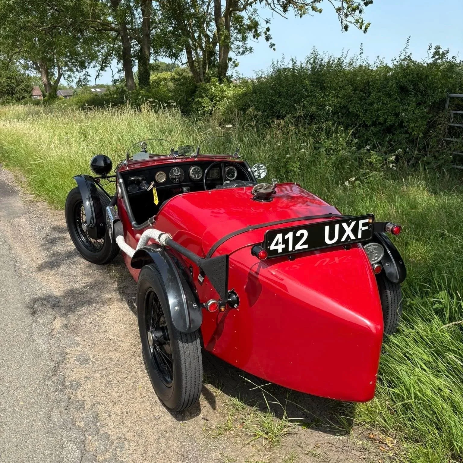 A vintage red three-wheeled race car with black wheels and a black dashboard, parked on a grassy roadside with trees and bushes in the background.