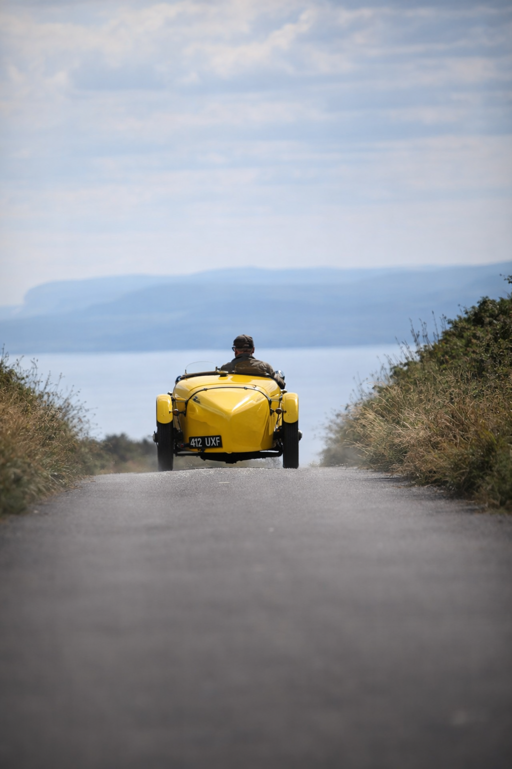 A person riding a yellow three-wheeled vehicle on a narrow road surrounded by bushes with a distant view of water and cliffs under a partly cloudy sky.