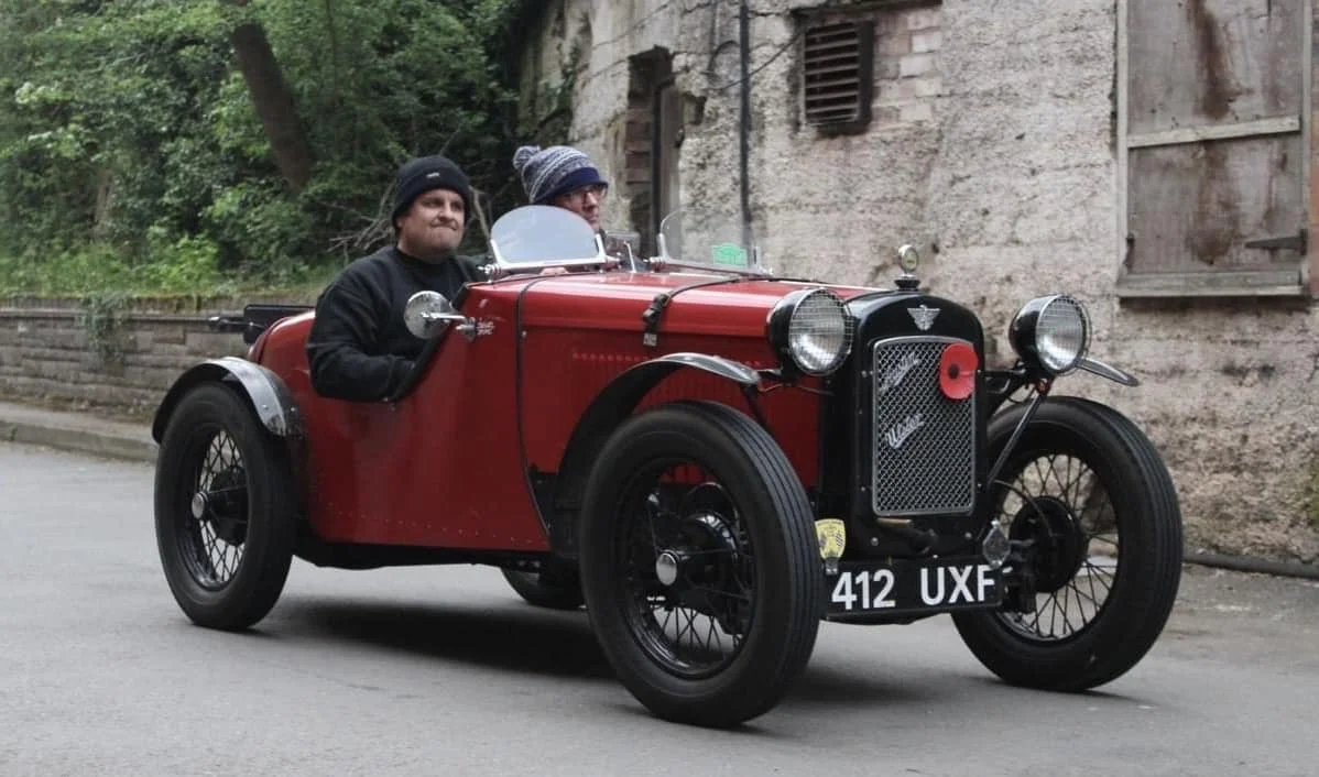 A red vintage open-top race car driven by two men in warm clothing and knit hats, driving along a street with green trees and an old stone building in the background.