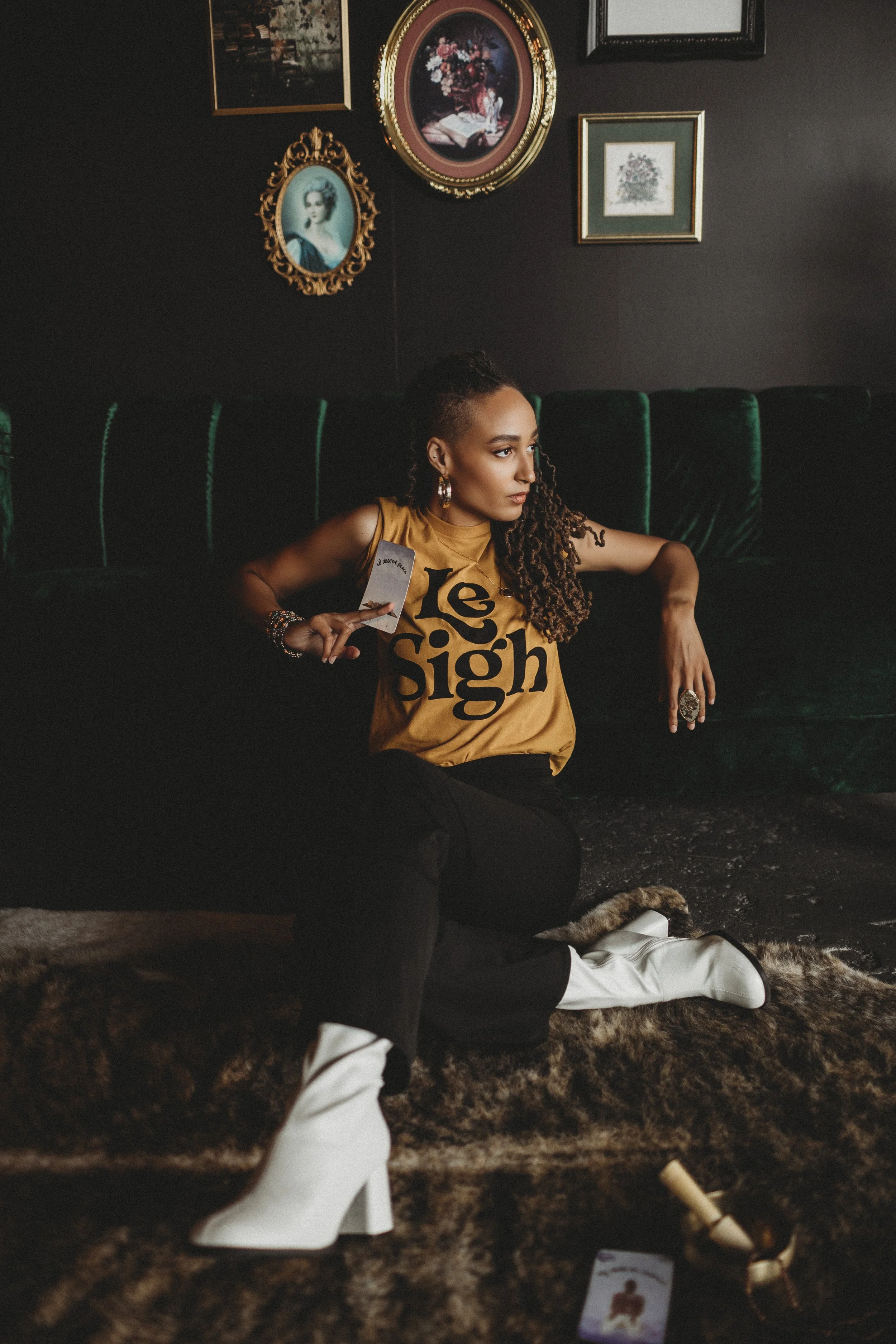 A woman sitting on a fur rug in a dimly lit room with dark walls decorated with framed artwork. She is wearing a mustard yellow shirt with 'Le Sigh' printed on it, black pants, and white high-heeled boots. She has long, curly hair, earrings, and rings, and is holding a card in her right hand. An earring or bracelet with a medallion design is visible on her left hand.