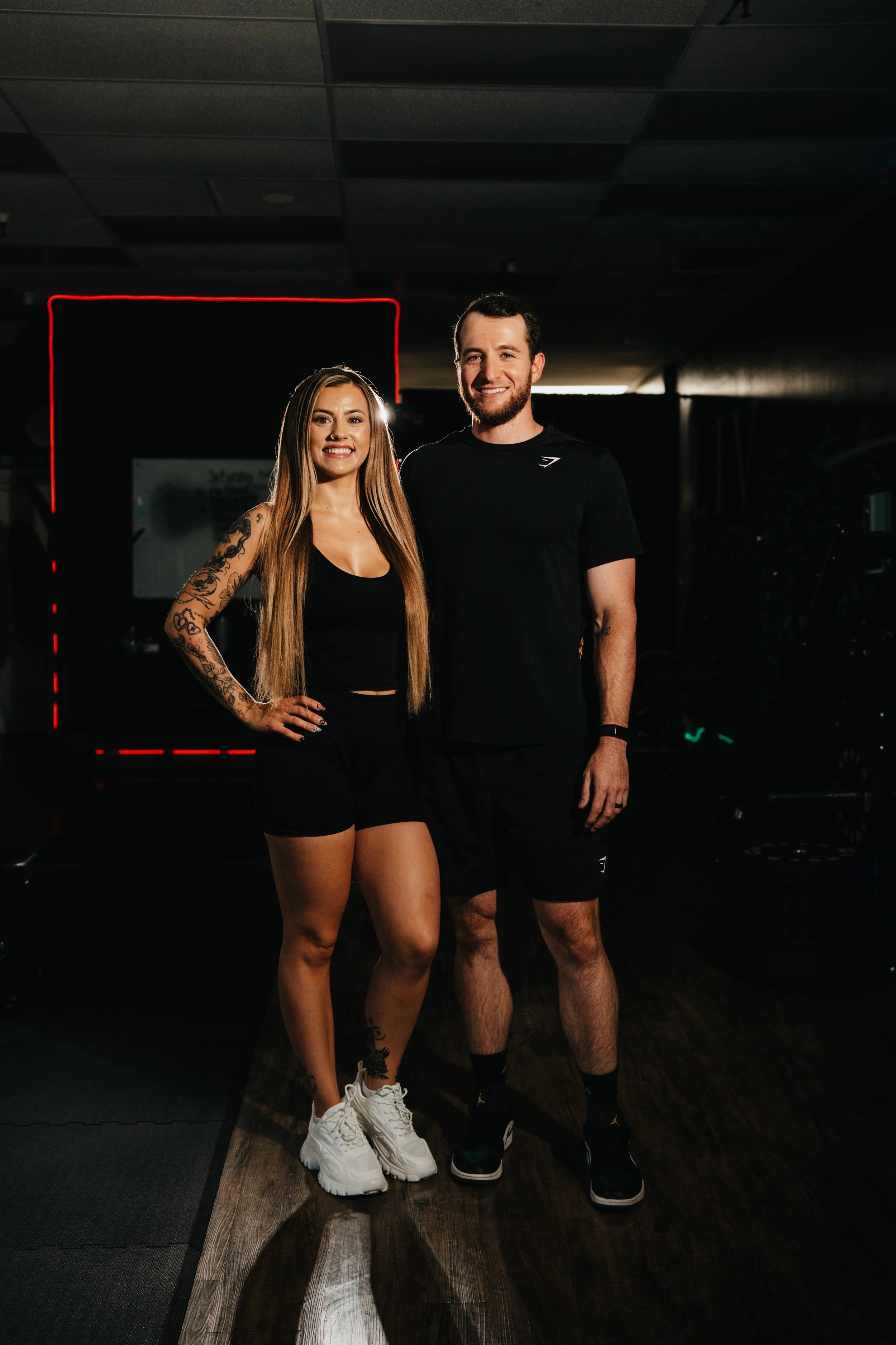 A smiling woman with long blonde hair and tattoos on her arms standing next to a smiling man with short dark hair and beard, both dressed in black clothing indoors against a light-colored wall.