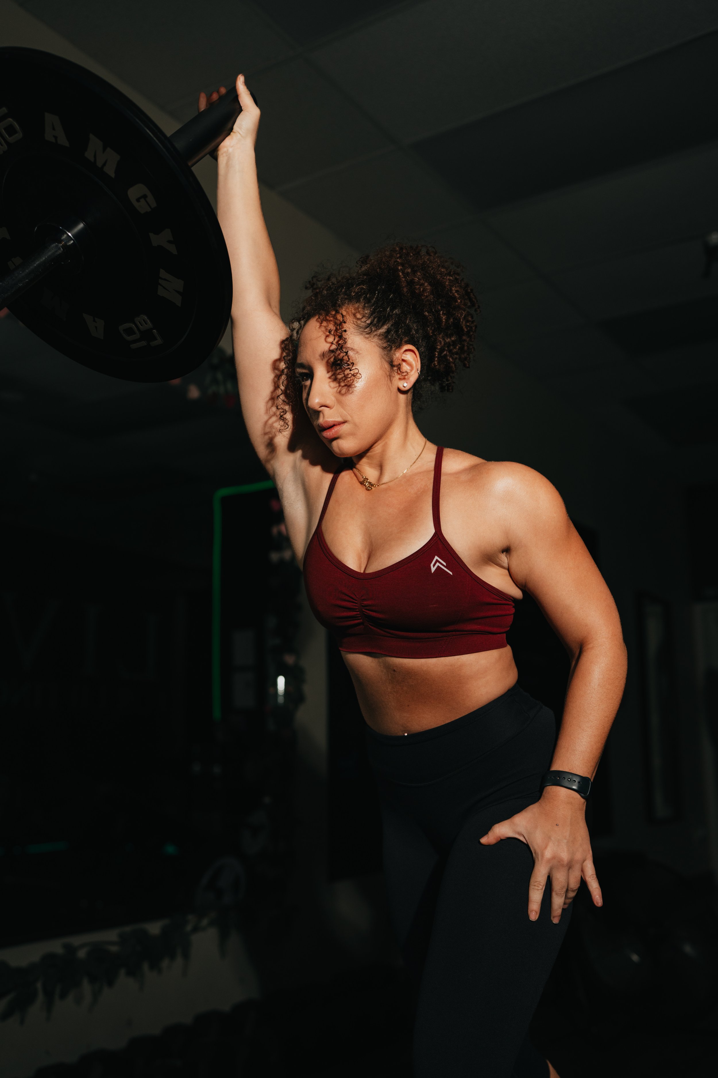 A smiling woman with curly hair in a gym, wearing a black sleeveless crop top with the words 'LIV FOR LIFTING' and black shorts, standing in front of gym equipment.