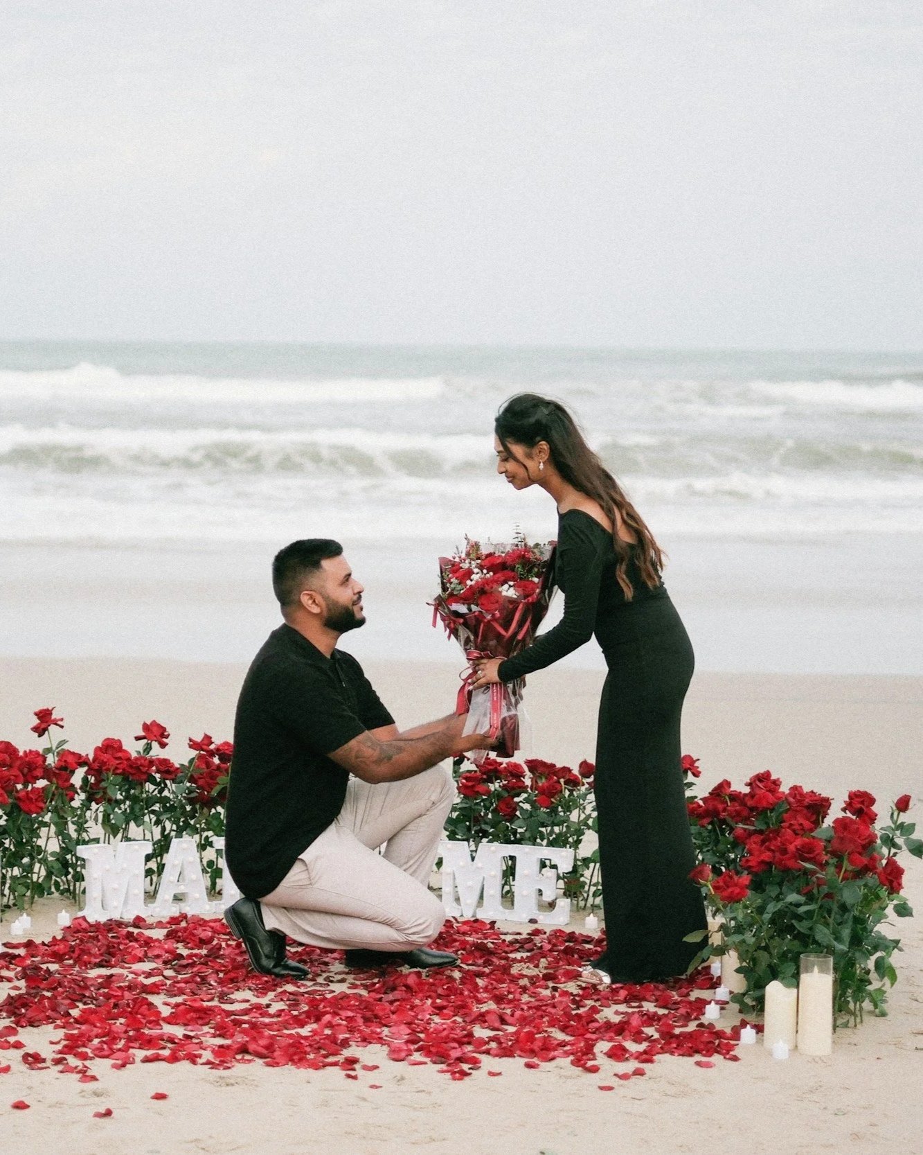 A man kneels on a sandy beach and proposes to a woman amid rose petals, flowers, and candles, with the ocean in the background.