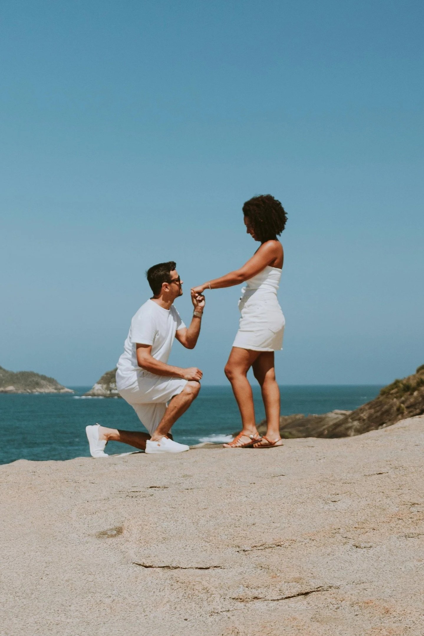 A man proposing marriage to a woman on the beach, with the man kneeling and holding the woman's hand against a backdrop of ocean and rocky islands under a clear blue sky.