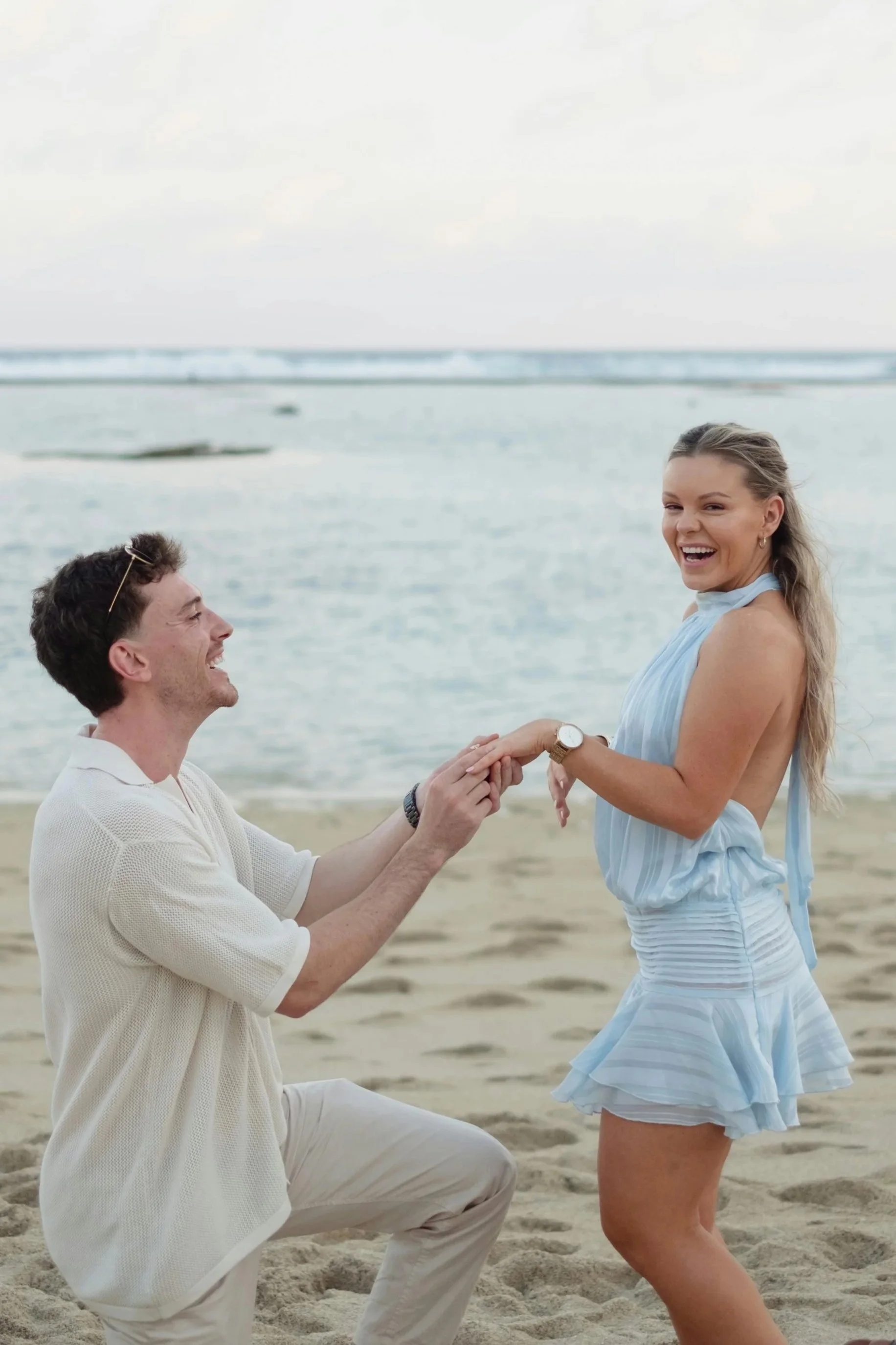 A man proposing marriage to a woman on the beach, with both smiling and holding hands, ocean in the background.